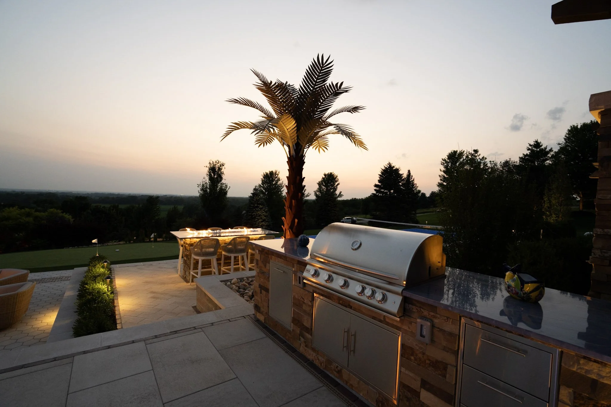 Outdoor patio with a grill, a table with chairs, a lit decorative plant, and a large artificial palm tree, overlooking a golf course and trees at sunset.