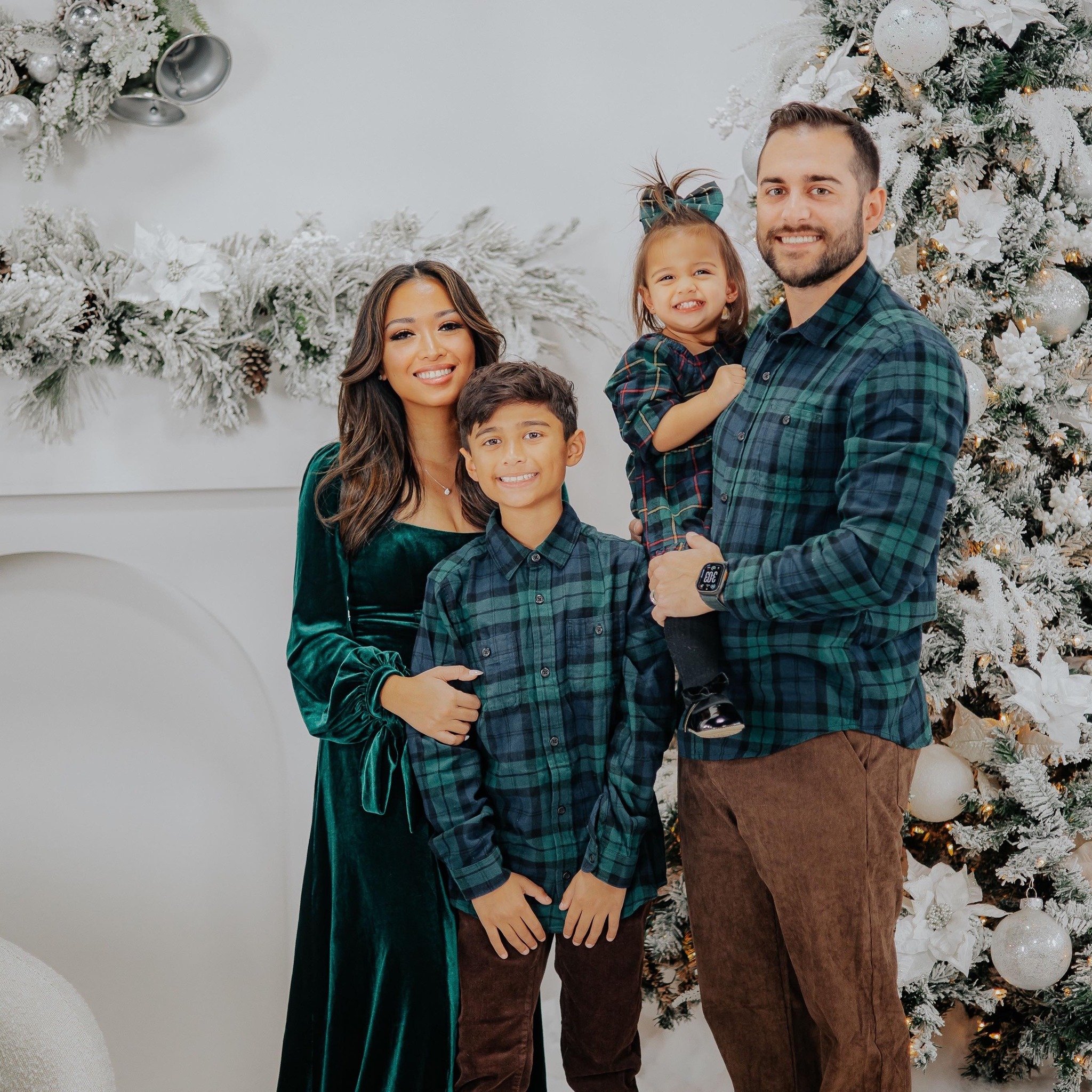 A family of four celebrating Christmas stands in front of a decorated Christmas tree and snowy garland with ornaments.