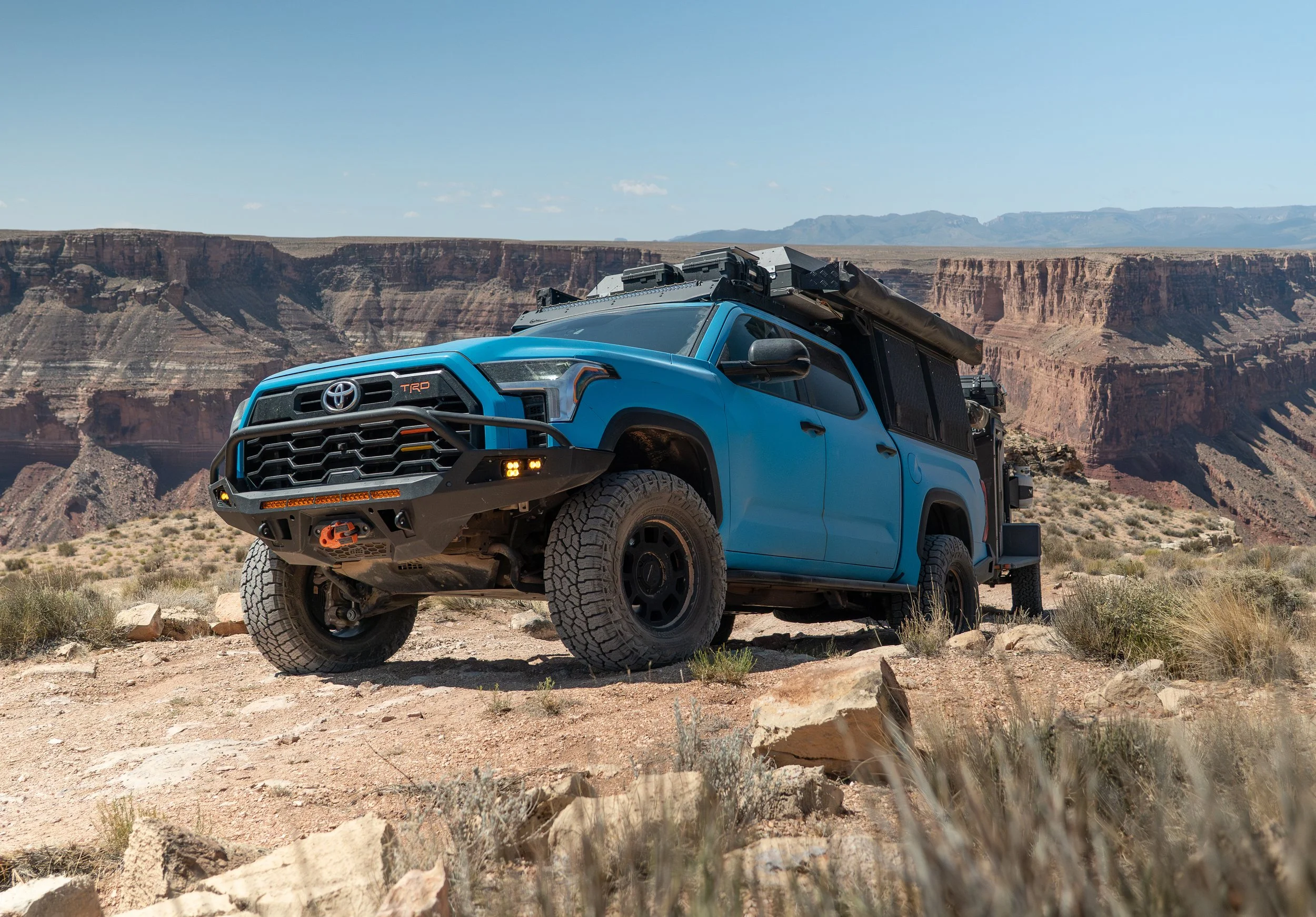 A blue off-road vehicle, possibly a Toyota Tacoma, equipped with gear on its roof and front bumper, parked in a desert landscape with canyon cliffs in the background.