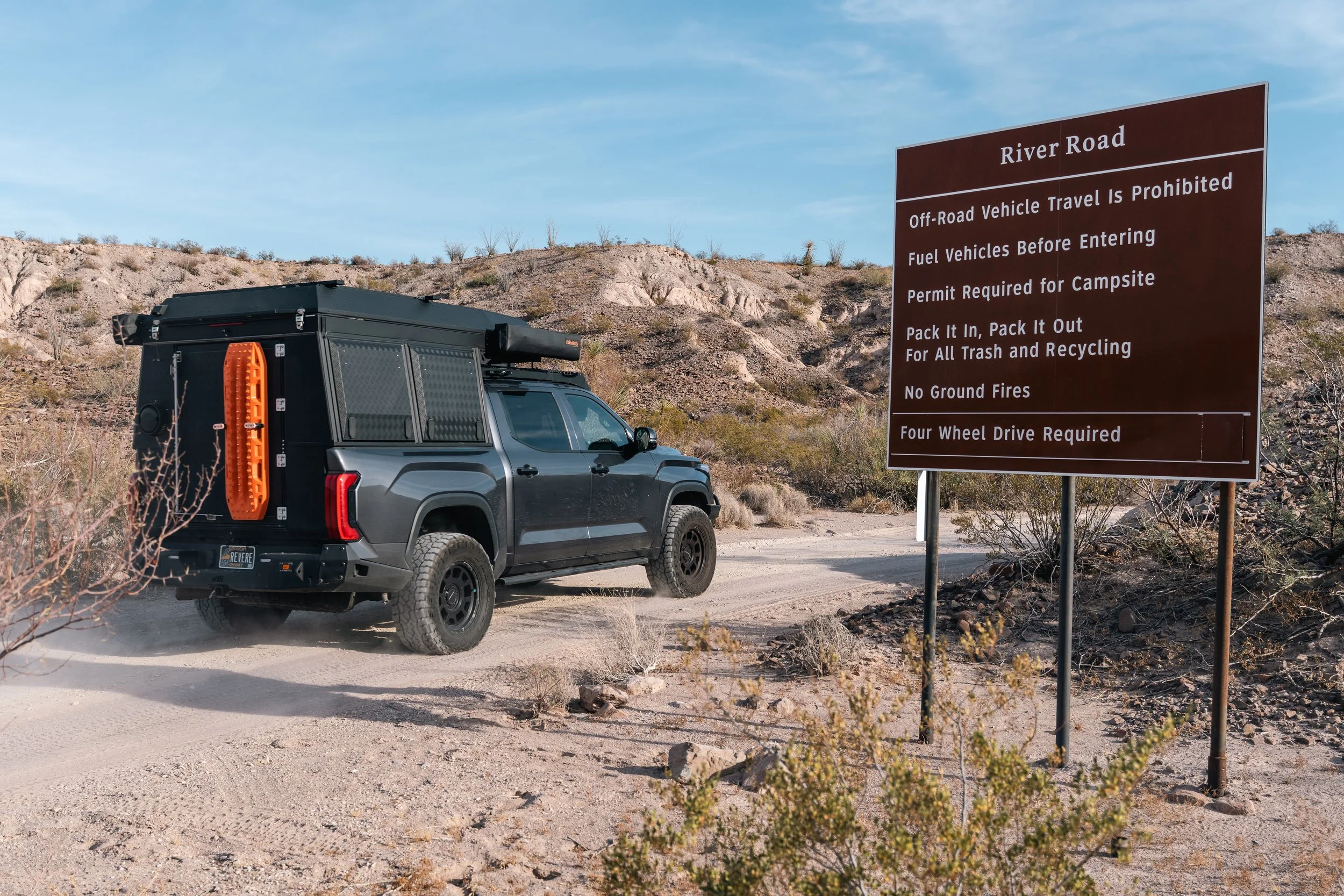 A black off-road vehicle parked on a dirt trail near a brown sign with white text at a desert area. The sign reads: River Road, Off-Road Vehicle Travel Is Prohibited, Fuel Vehicles Before Entering, Permit Required for Campsite, Pack It In, Pack It Out for All Trash and Recycling, No Ground Fires, Four Wheel Drive Required.