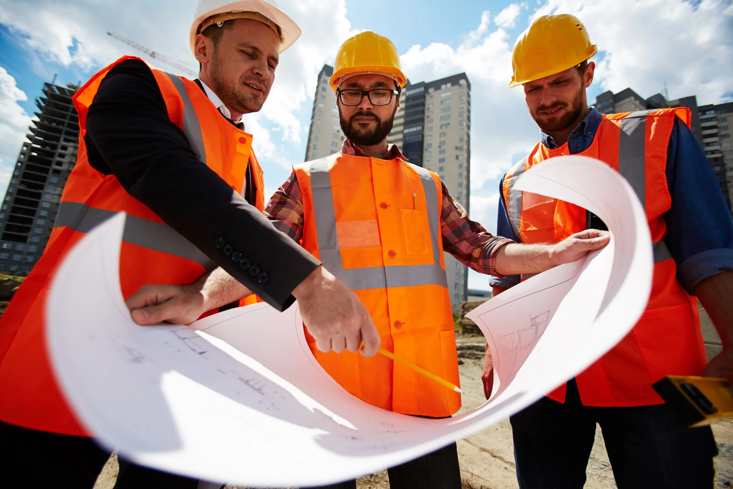 Three male construction workers wearing yellow hard hats and orange safety vests examining blueprints at a construction site.