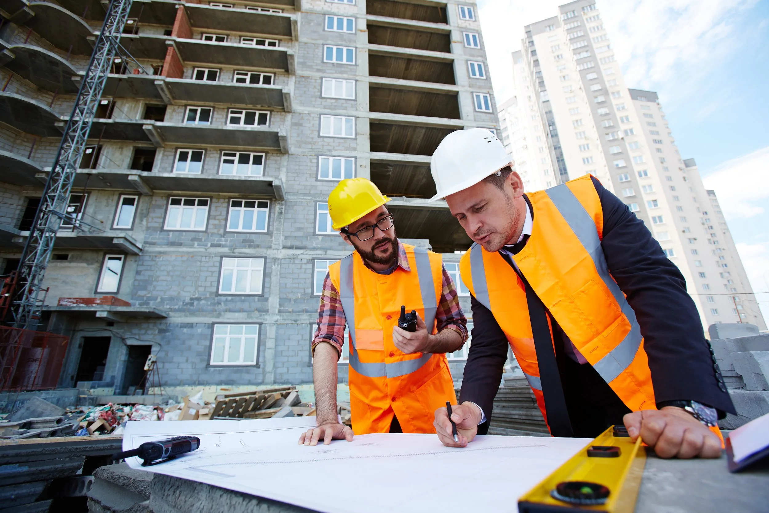 Two construction workers in safety vests and helmets reviewing blueprints at a building site with an unfinished high-rise building in the background.