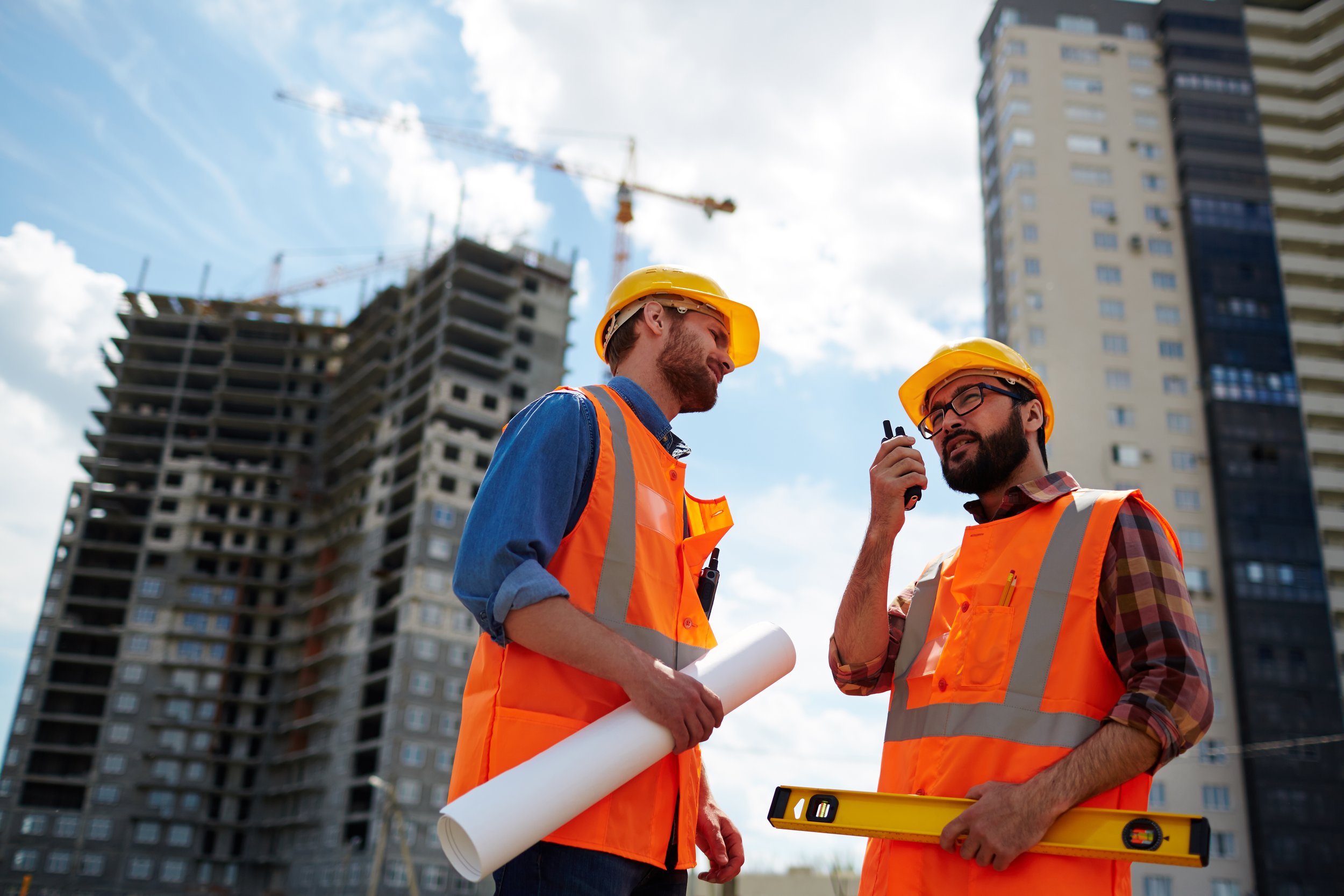 Two construction workers in orange safety vests and helmets discussing on a construction site with a high-rise building under construction in the background.