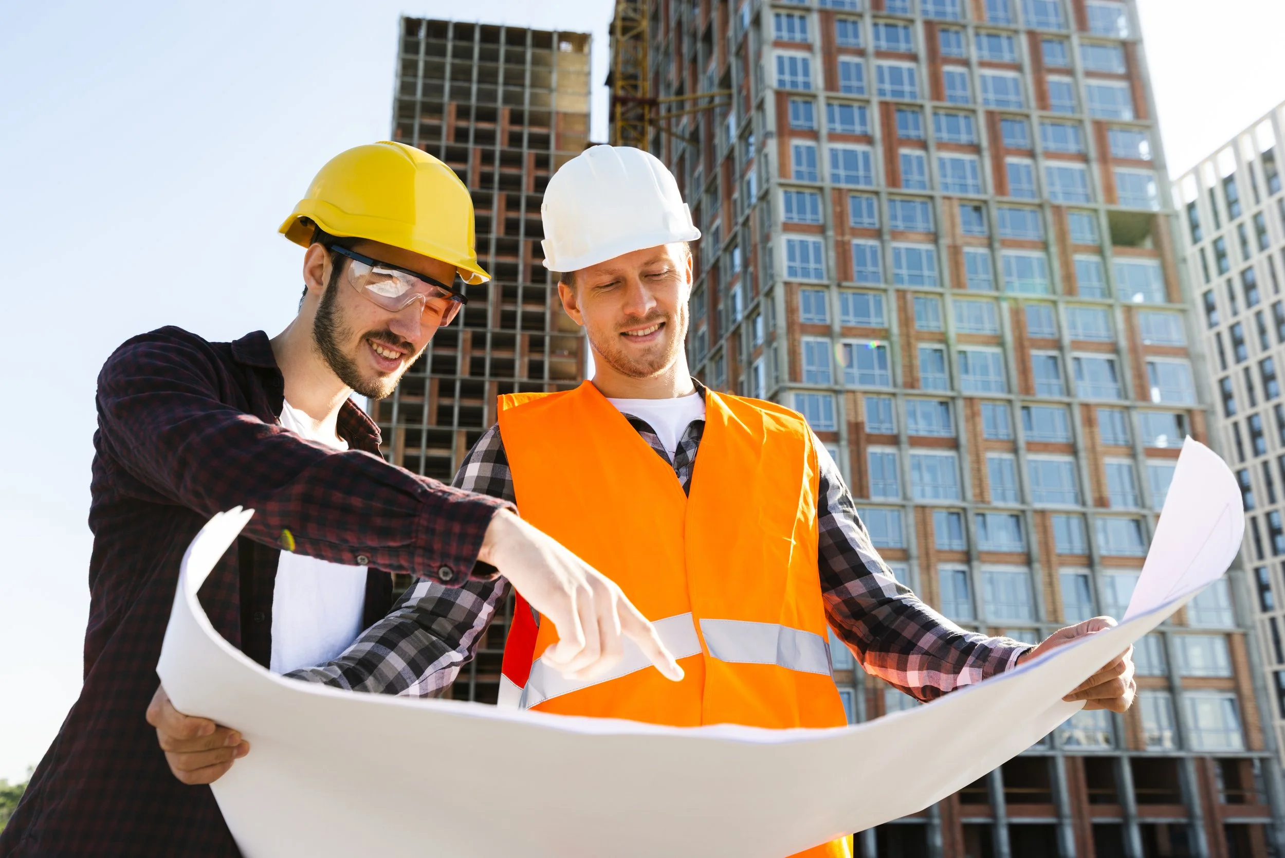 Two construction workers, one wearing a yellow hard hat and safety goggles and the other wearing a white hard hat and an orange safety vest, look at a blueprint at a construction site with high-rise buildings in the background.