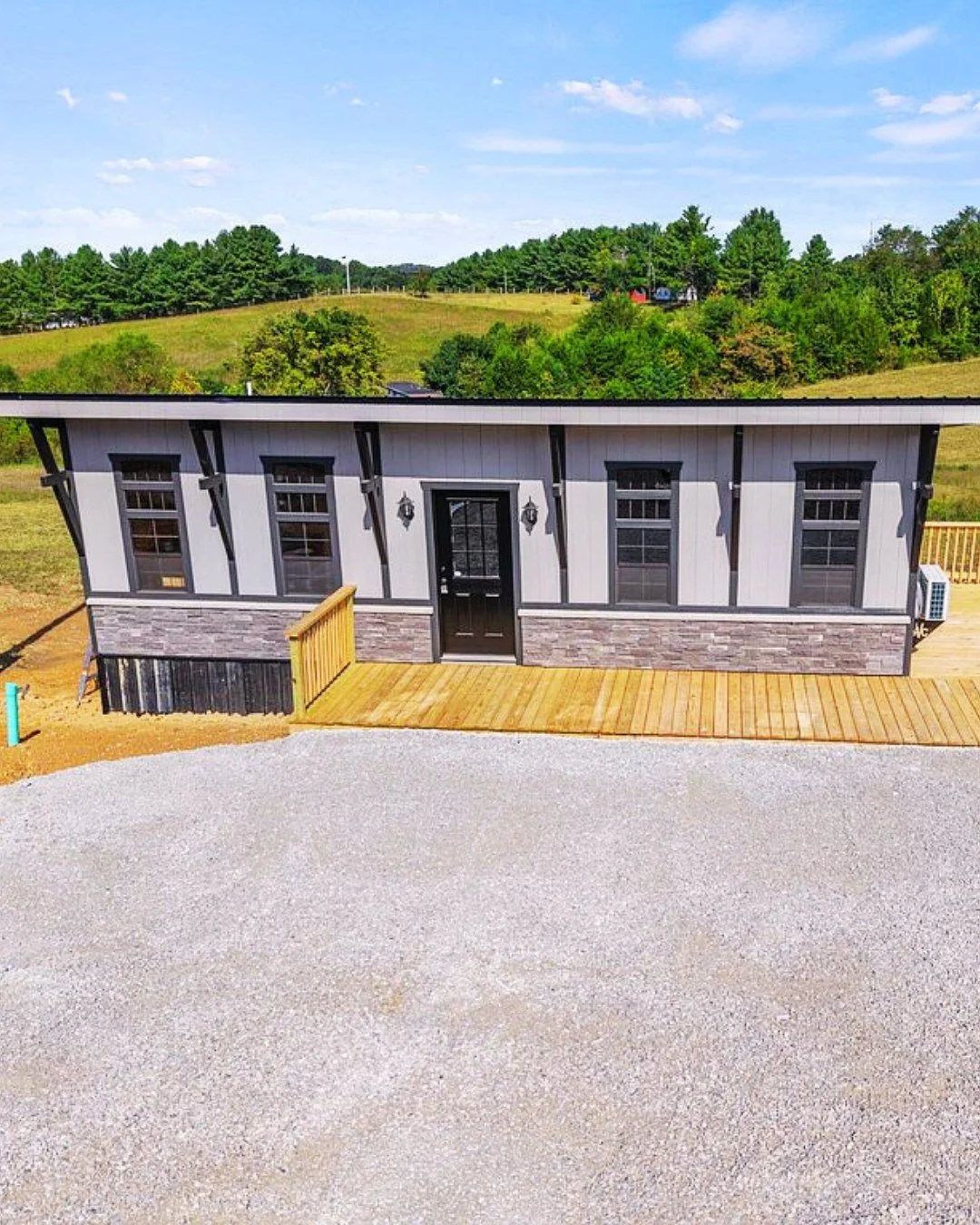 A small modern house with gray siding, black trim, and a brick half-wall along the bottom, situated on a wooden deck with a gravel driveway in front, surrounded by green hills and trees in the background.