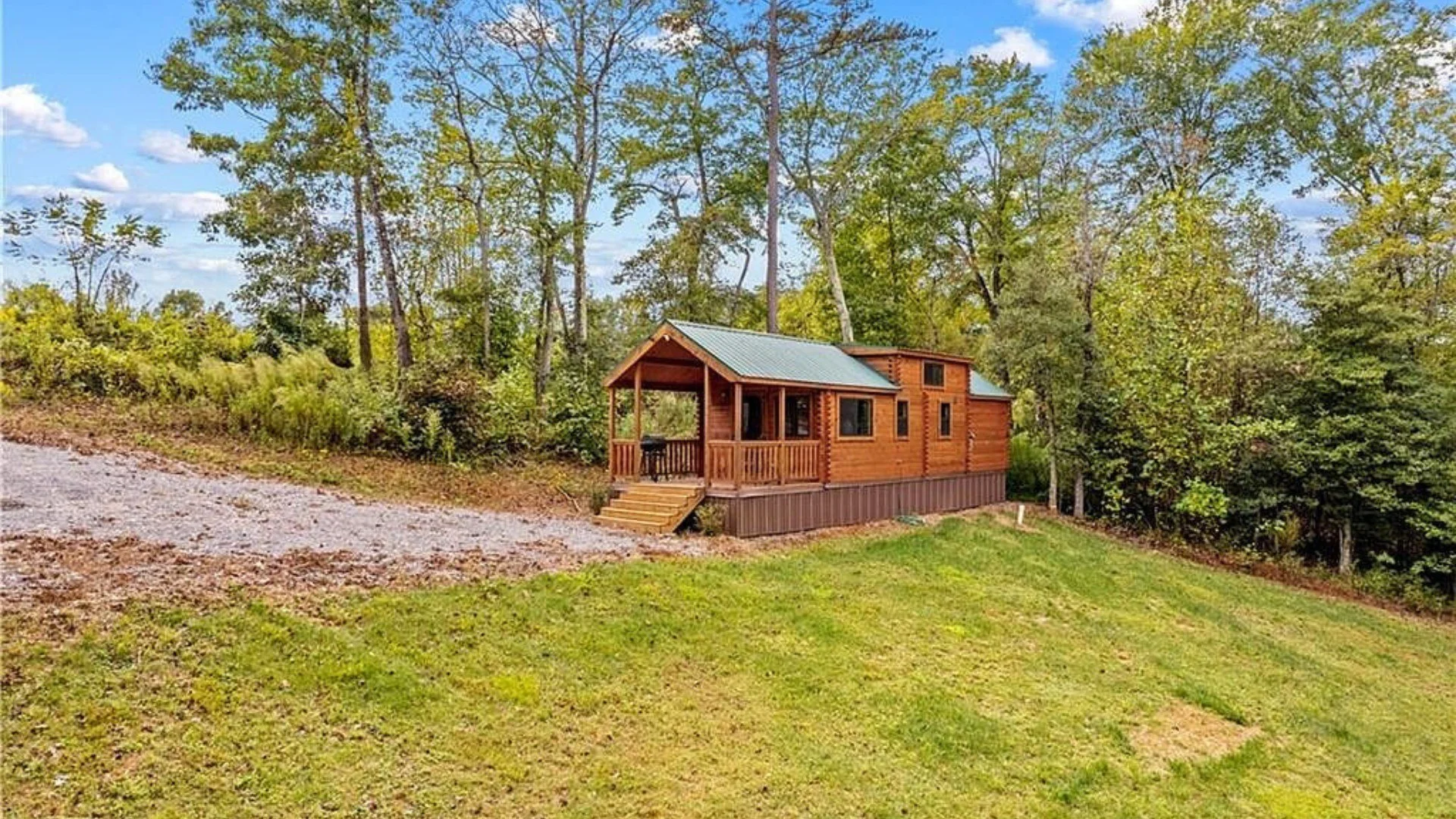A small wooden cabin with a porch and metal roof, situated on a hillside amid trees with green foliage, under a partly cloudy sky.