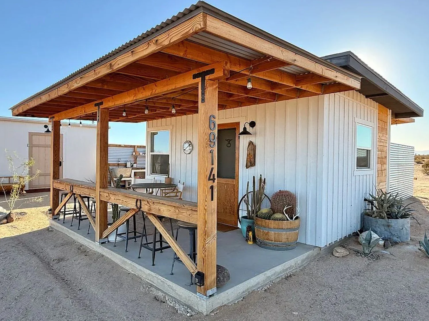 Small modern house with a wooden porch, outdoor seating, potted cacti, and white siding in a desert area.