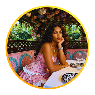 A woman sitting at a beautifully set outdoor table with floral decoration in the background, wearing a pink dress and jewelry, with empty wine glasses and black and white plates.
