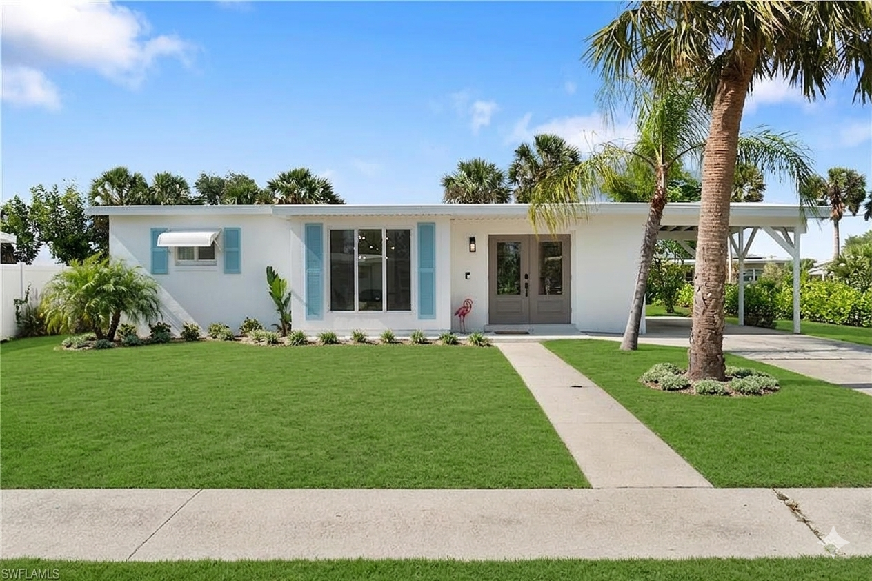 Front view of a modern, single-story house with white walls, blue shutters, and a gray front door, surrounded by a lush green lawn, palm trees, and tropical plants, under a clear blue sky.