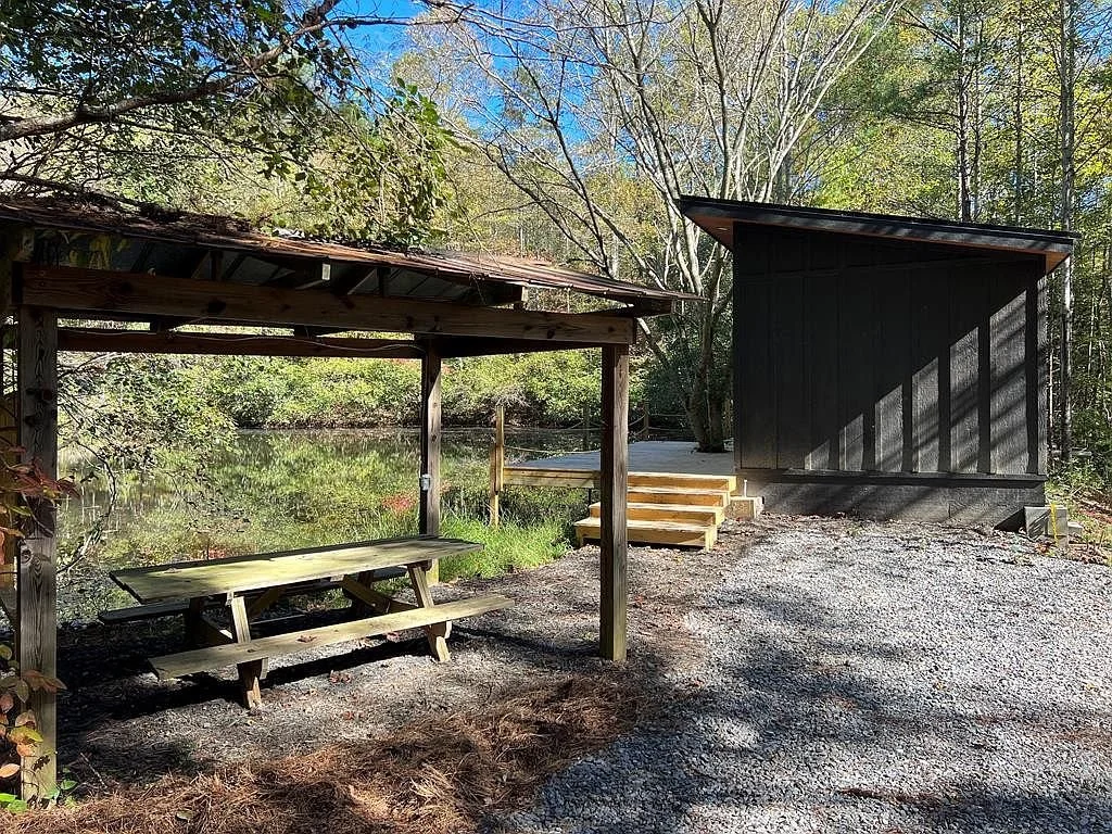 A small lakeside shelter with a picnic table underneath, adjacent to a black wooden building, with wooden stairs leading up to a dock near trees and water.