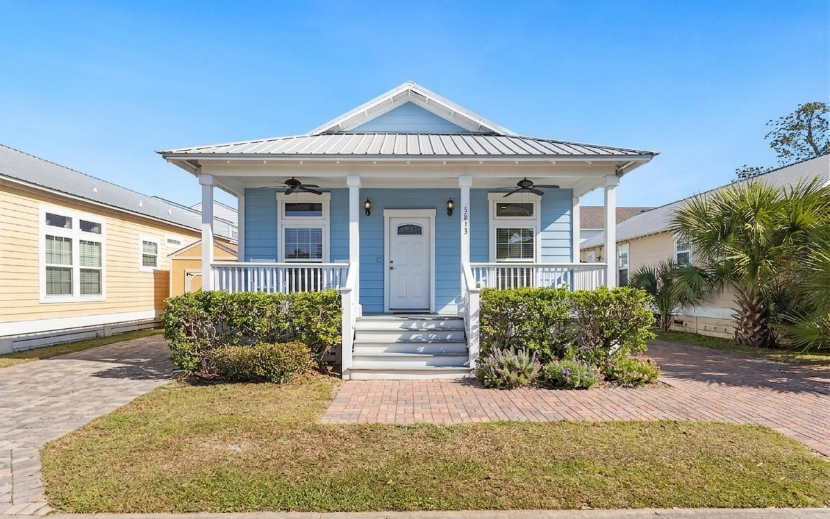 Front view of a blue house with a white door, front porch, and small steps, surrounded by green bushes and palm trees, under a clear blue sky.