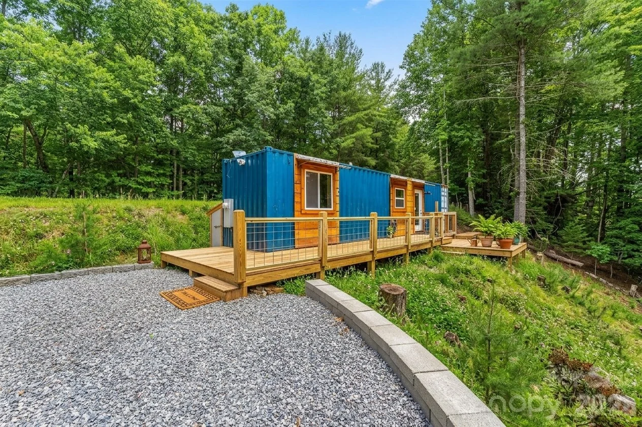 A small blue shipping container converted into a tiny home, positioned on a wooden deck with a gravel pathway and surrounded by a green forested area.