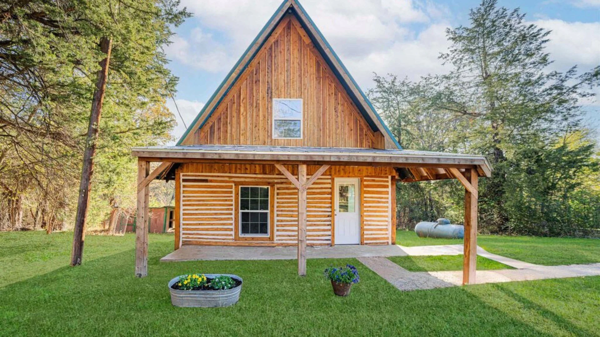 Wooden house with a steep roof and a front porch, surrounded by green grass and trees, with flower pots and a propane tank nearby.