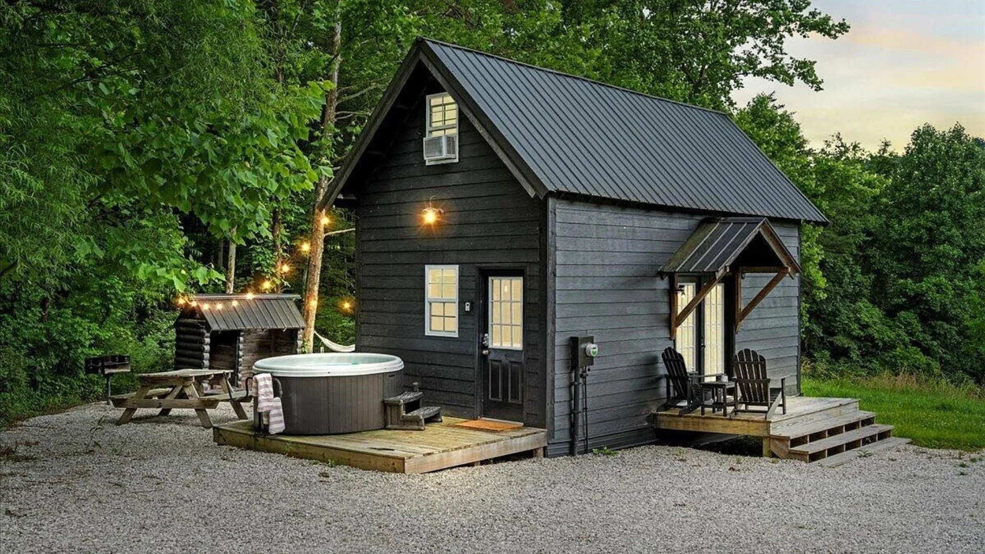 A small black wooden cabin with a metal roof, surrounded by trees. It features a porch with chairs, string lights hanging from trees, a hot tub on a deck, and a small cabin with a doghouse in the yard. Tall trees with green leaves are in the background, and the sky is slightly overcast.