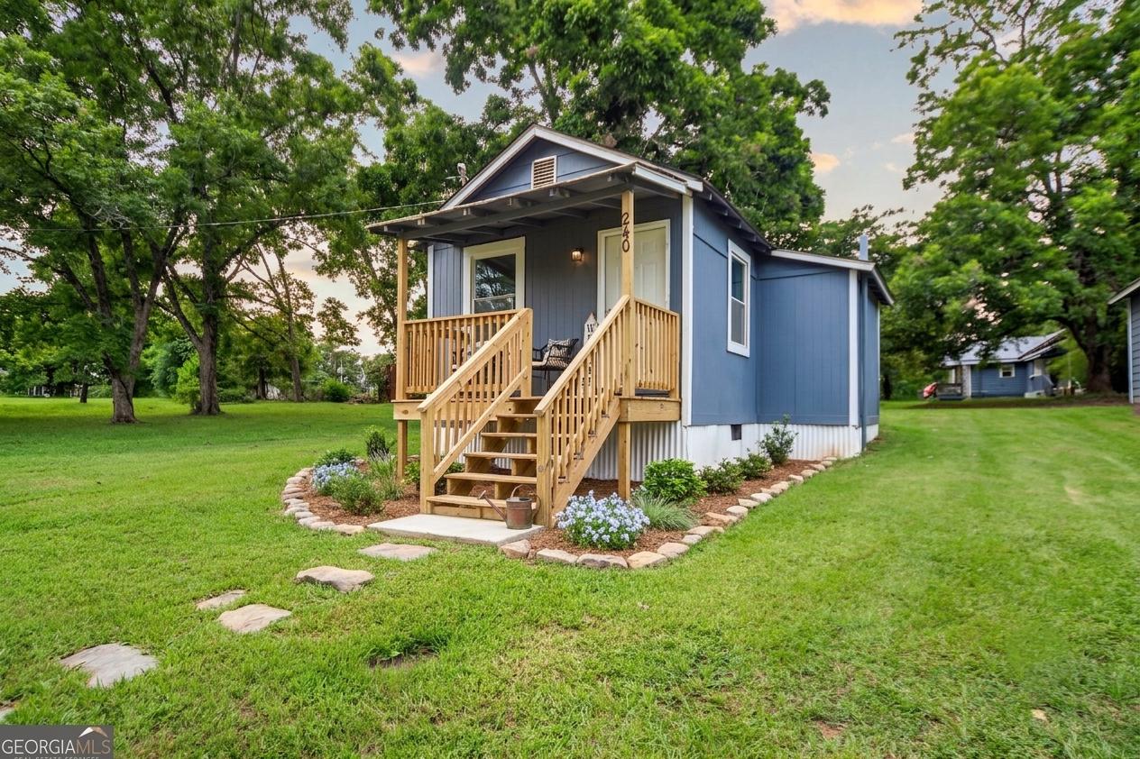 Front view of a blue house with a small front porch and wooden stairs, surrounded by a neatly landscaped lawn with garden beds, flowers, and trees.