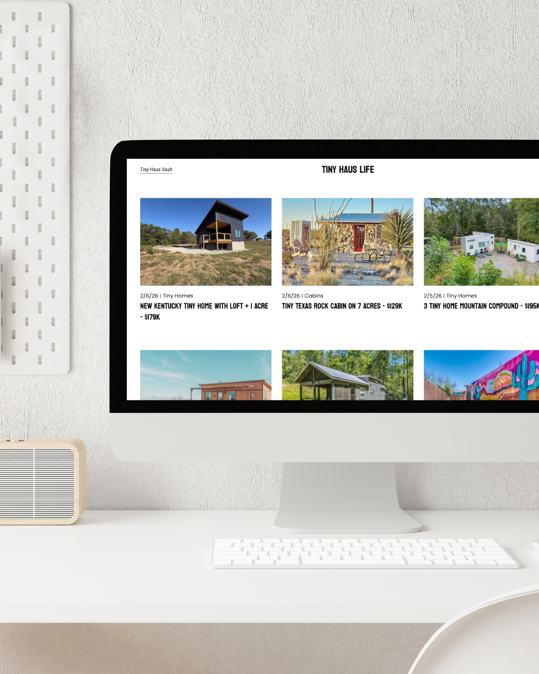 Computer monitor displaying a webpage with listings for tiny homes and cabins, set on a white desk with a white keyboard, a speaker, and a perforated white panel to the side.