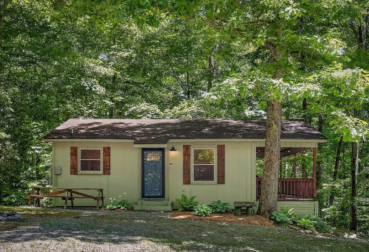A small house with green exterior walls, wooden shutters on the windows, and a black front door with a small set of steps leading up to it. The house is surrounded by trees and greenery, with a porch on the right side and a bench in front.