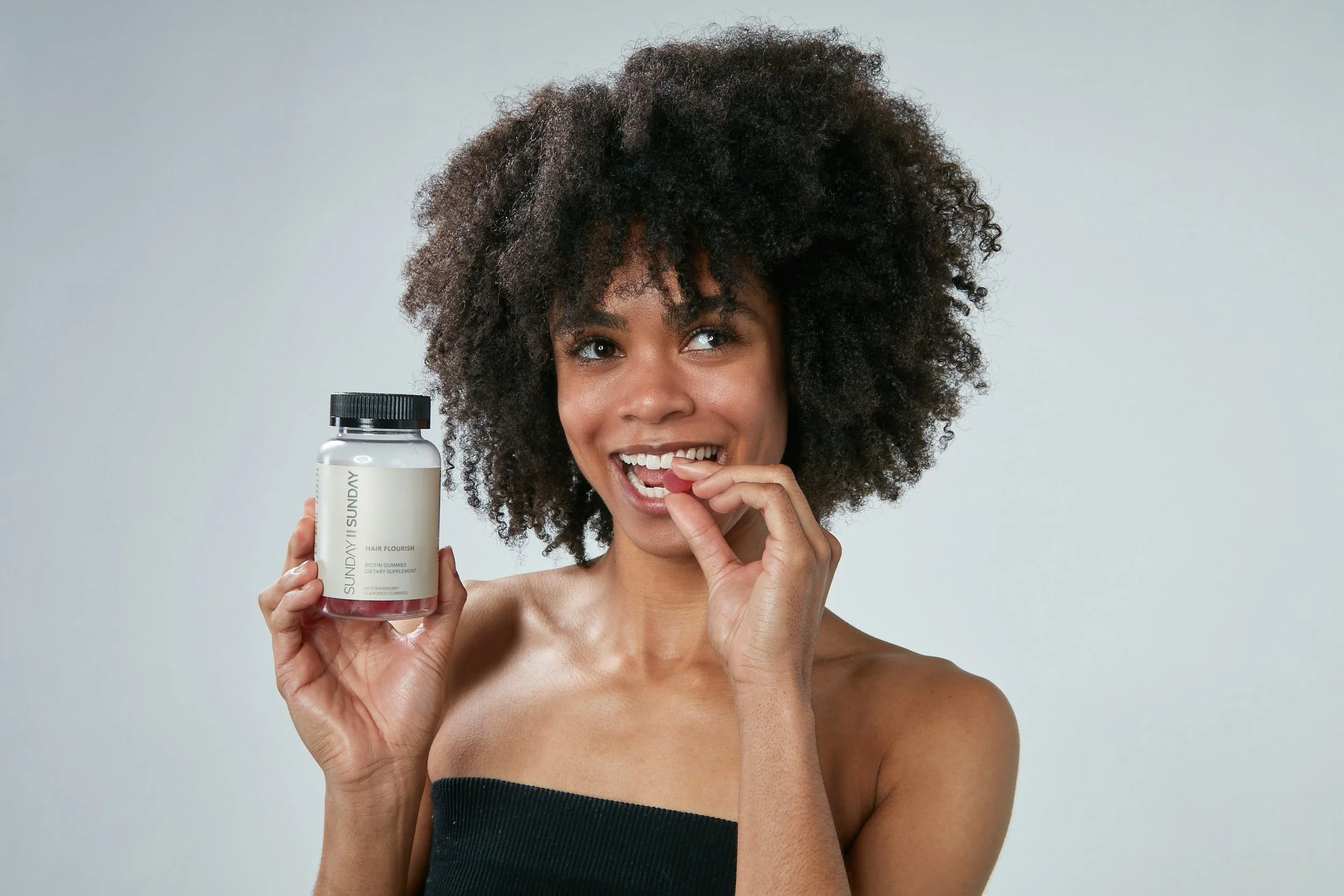 A woman with natural curly hair holding a bottle of hair supplement and smiling.