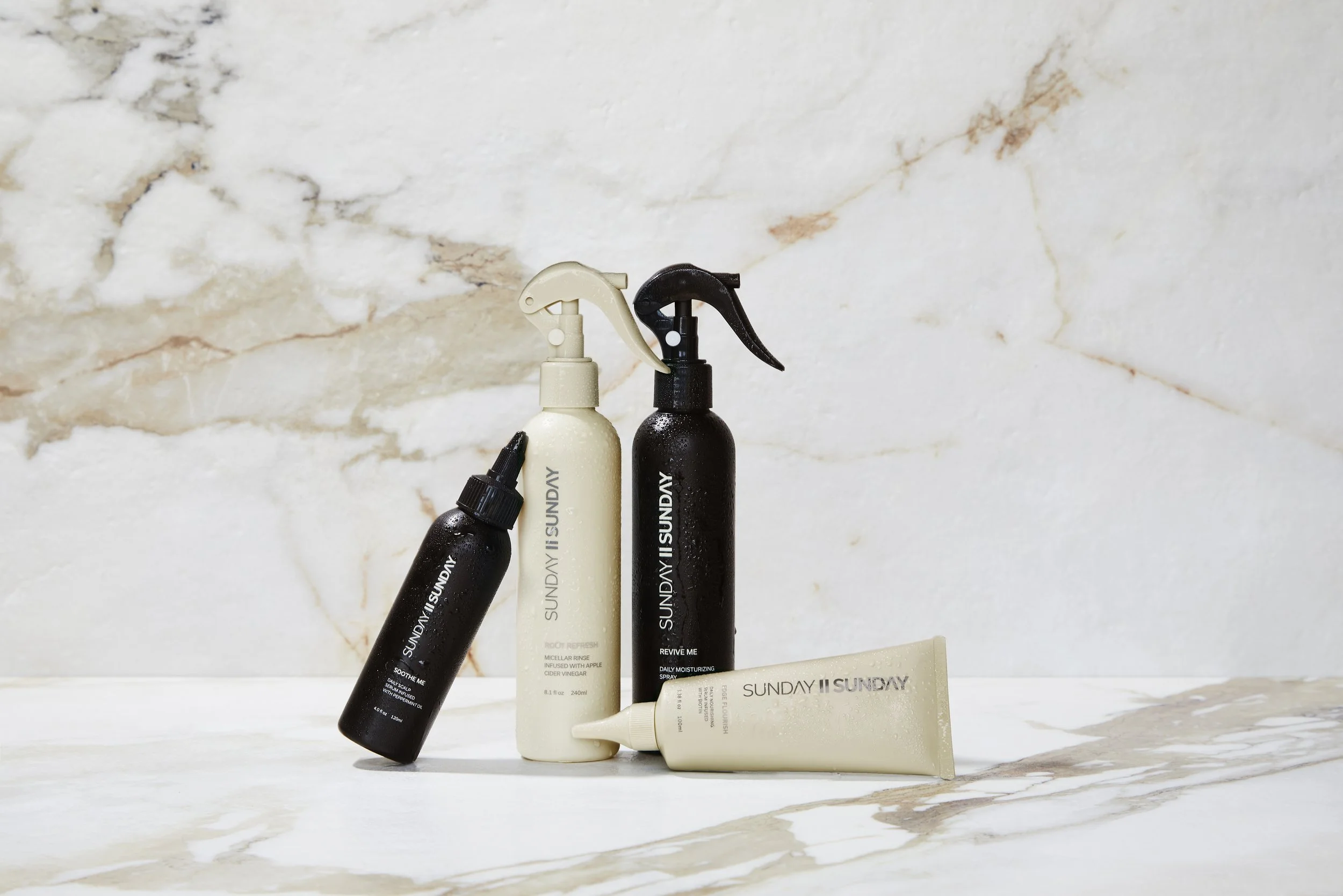 Collection of four skincare bottles and tubes on marble surface with marble wall background, some bottles have spray nozzles and one has a dropper cap.