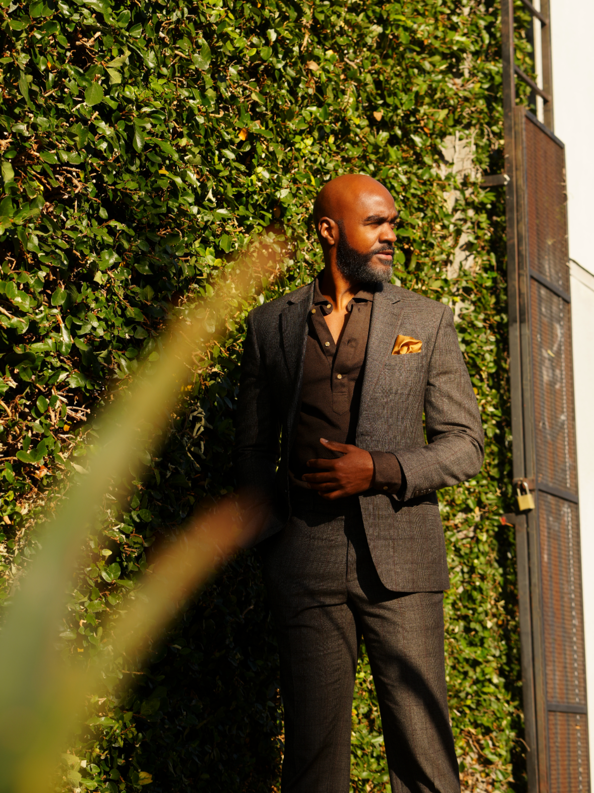 Myron Branford in a brown suit with a gold pocket square stands against a lush green hedge, looking to the side with sunlight illuminating his face.