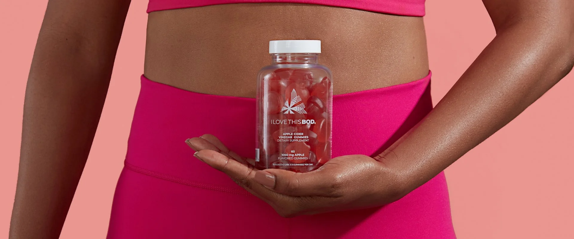 A woman wearing pink athletic wear holds a clear bottle of gummy vitamins labeled 'I Love This BOD' with apple cider vinegar gummies inside, against a pink background.