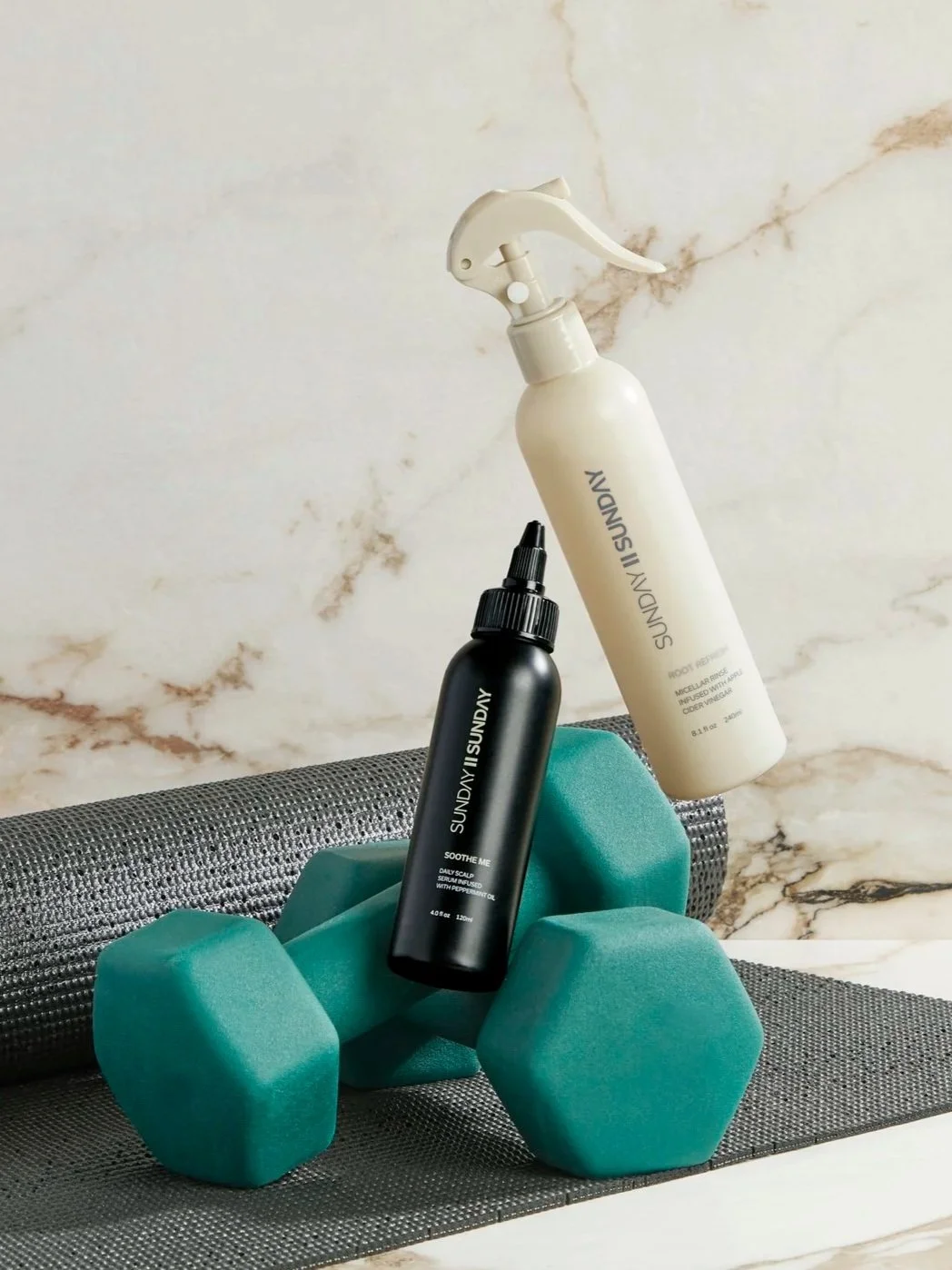 Workout and skincare items on a yoga mat, including a black bottle labeled 'Soothe Me' with a spray nozzle, a white spray bottle labeled 'SunDay SunDay,' and a gray foam roller, with teal dumbbells and a marble background.