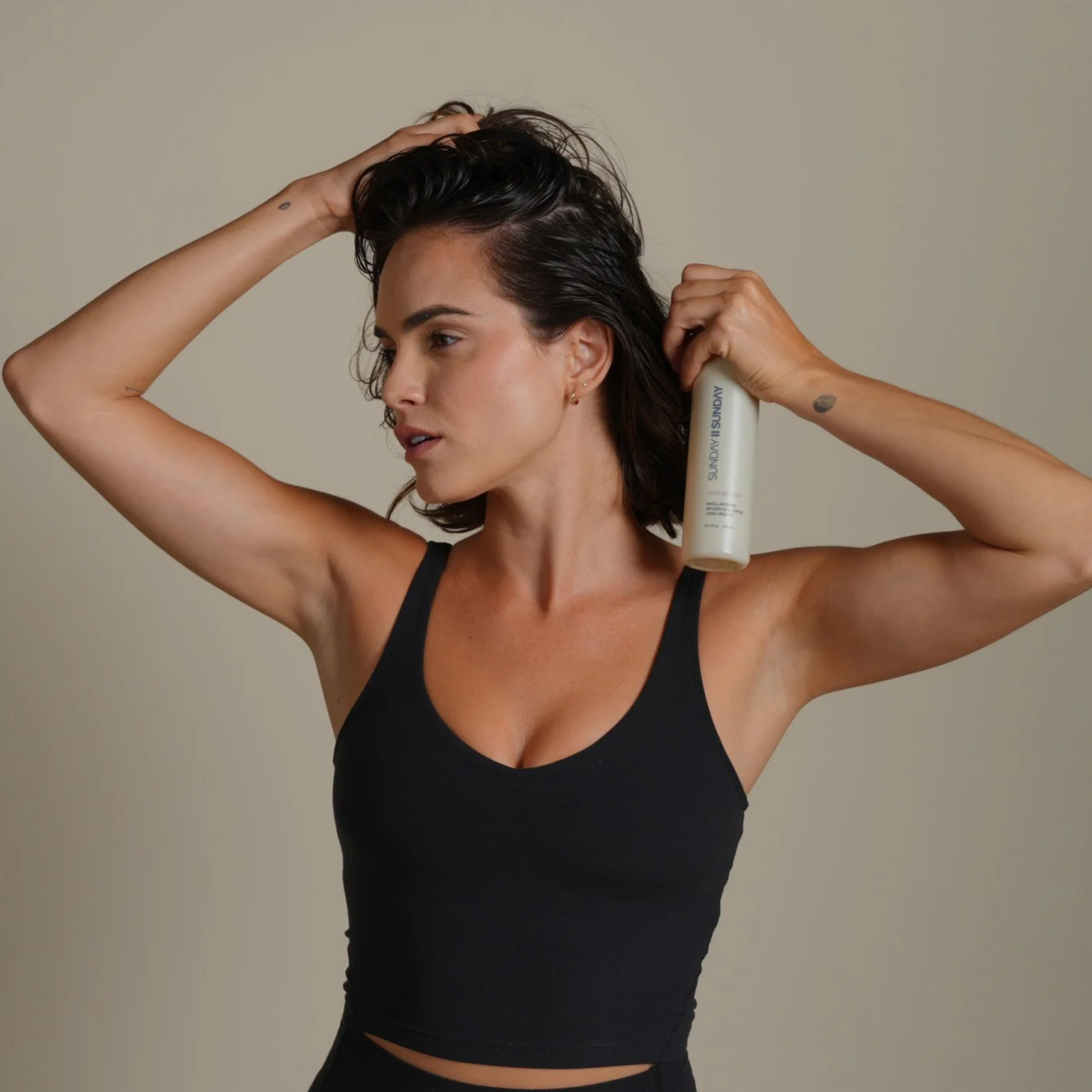 Woman with short dark hair in a black tank top applying hair product from a beige spray bottle against a plain beige background.