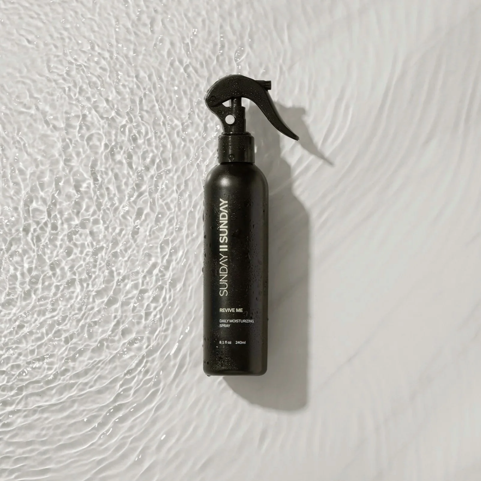 A black spray bottle labeled 'SUNDY II SUNDAY' with the words 'REVIVE ME DAILY MOISTURIZING SPRAY' on a textured white surface.