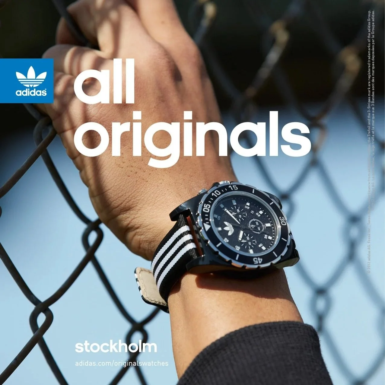 A close-up of a person's wrist wearing an Adidas watch with a black and white striped strap, holding onto a chain-link fence with a blue sky in the background.