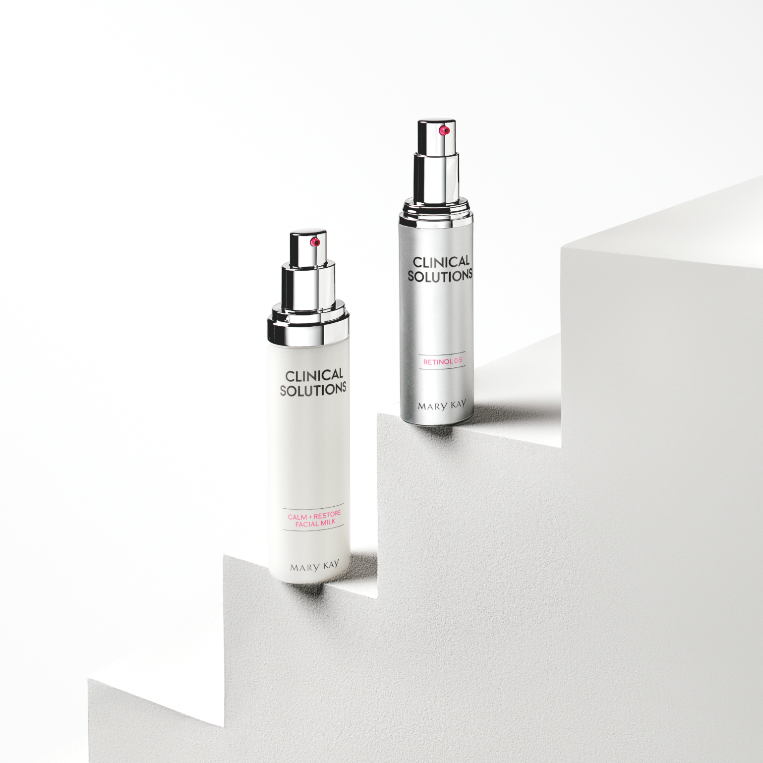 Two skincare bottles labeled 'Clinical Solutions' by Mary Kay, one labeled 'Calm + Restore Facial Milk' and the other 'Retinol 0.5', positioned on a white stepped surface with a plain white background.