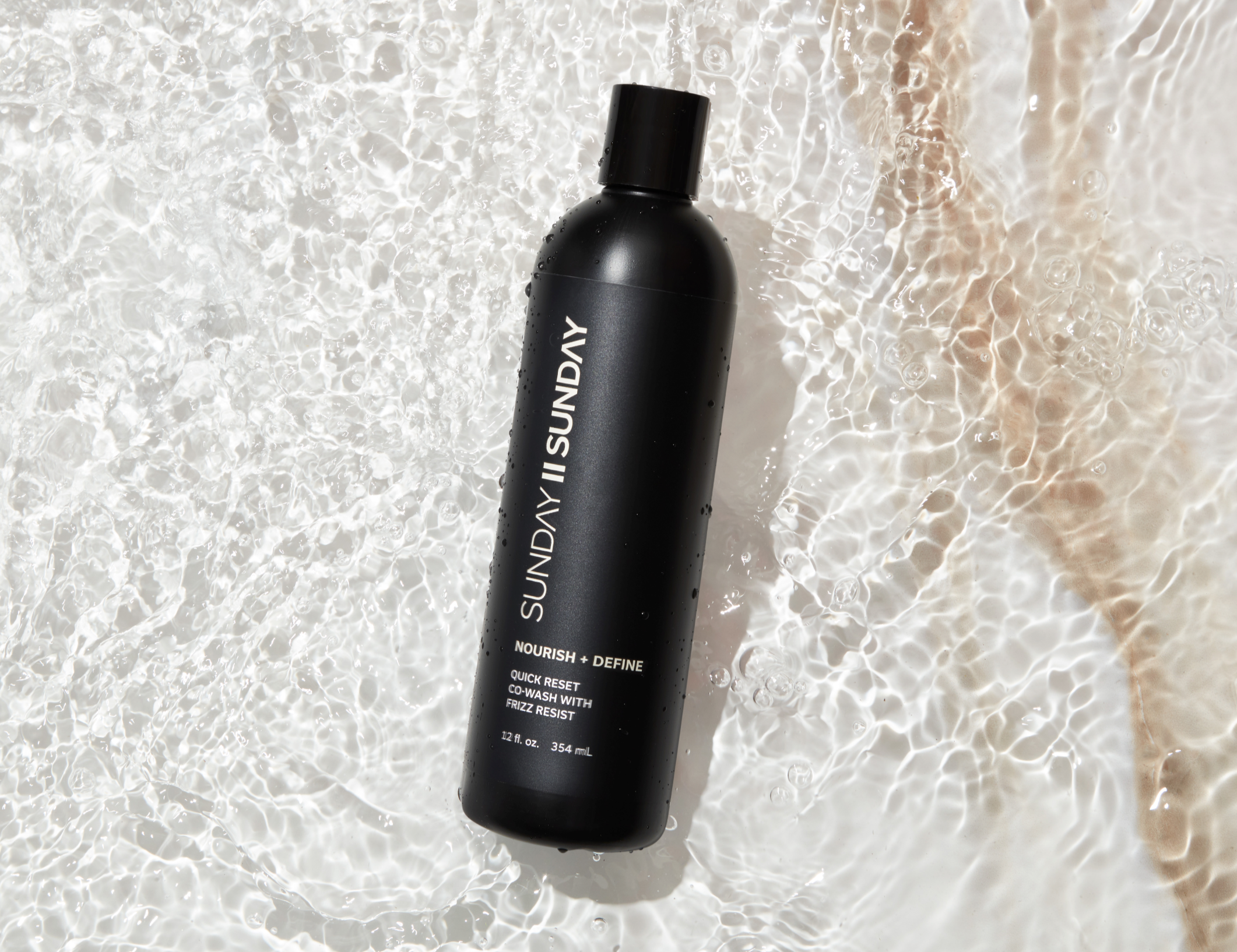 Black bottle of shampoo or conditioner labeled "SUNDAY II SUNDAY" lying on a sunlit water surface with ripples and bubbles.