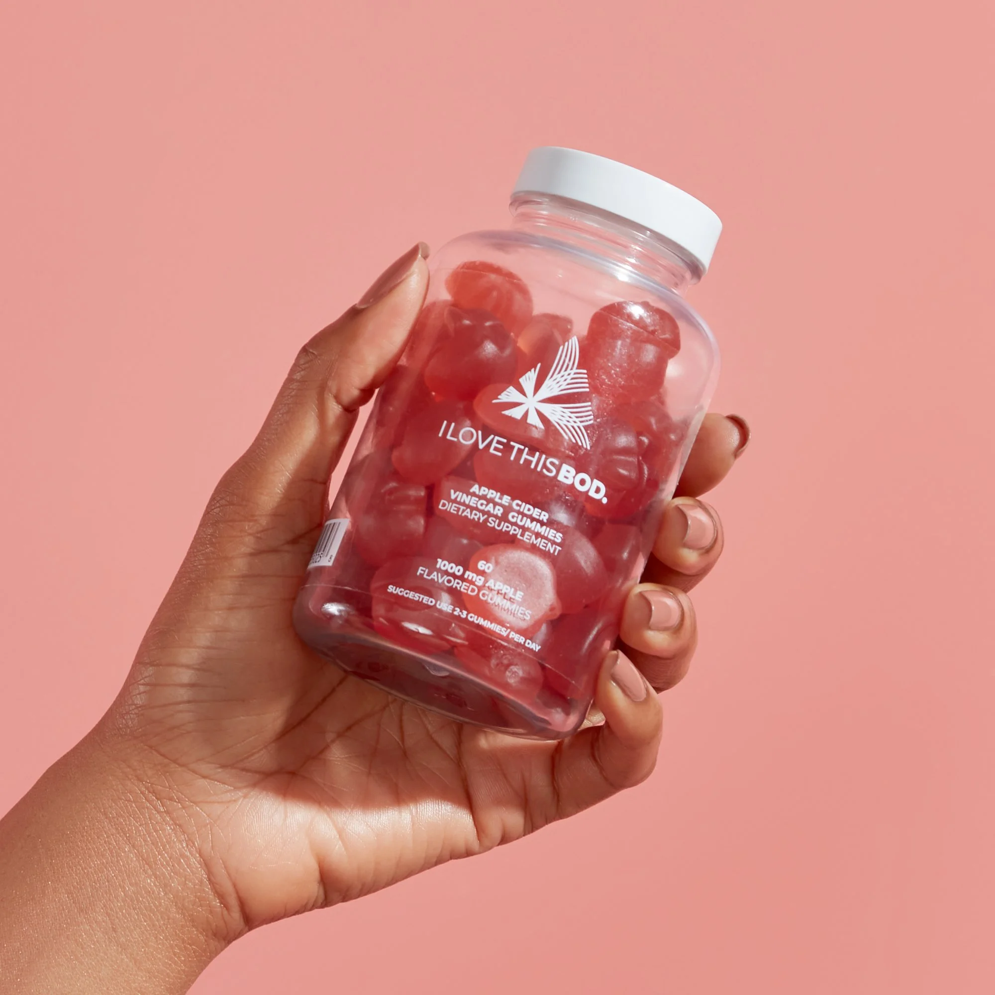 Hand holding a clear jar of red apple cider vinegar gummies with a white lid, set against a pink background.