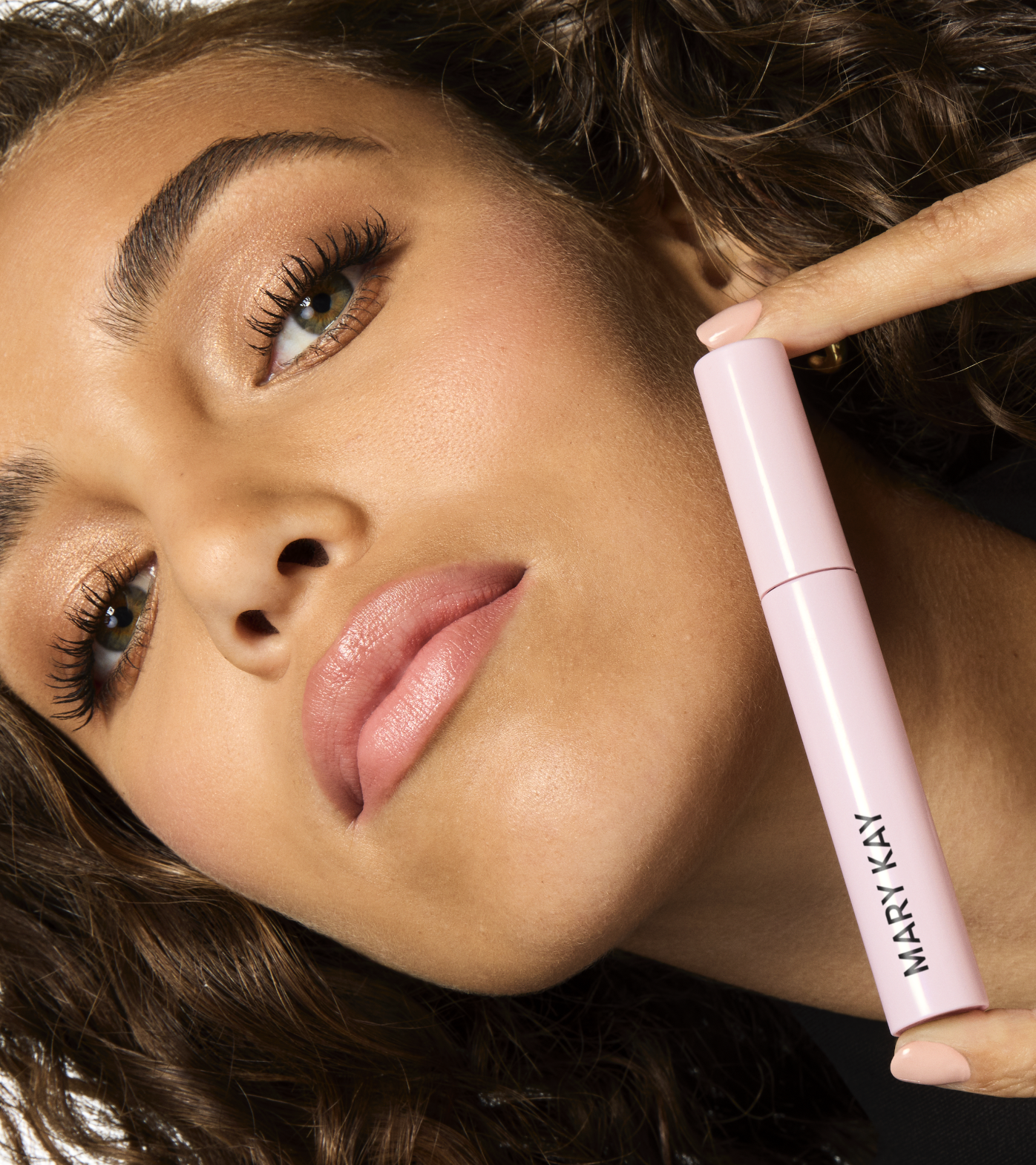 Close-up of a woman with curly brown hair holding a pink Mary Kay product near her face.