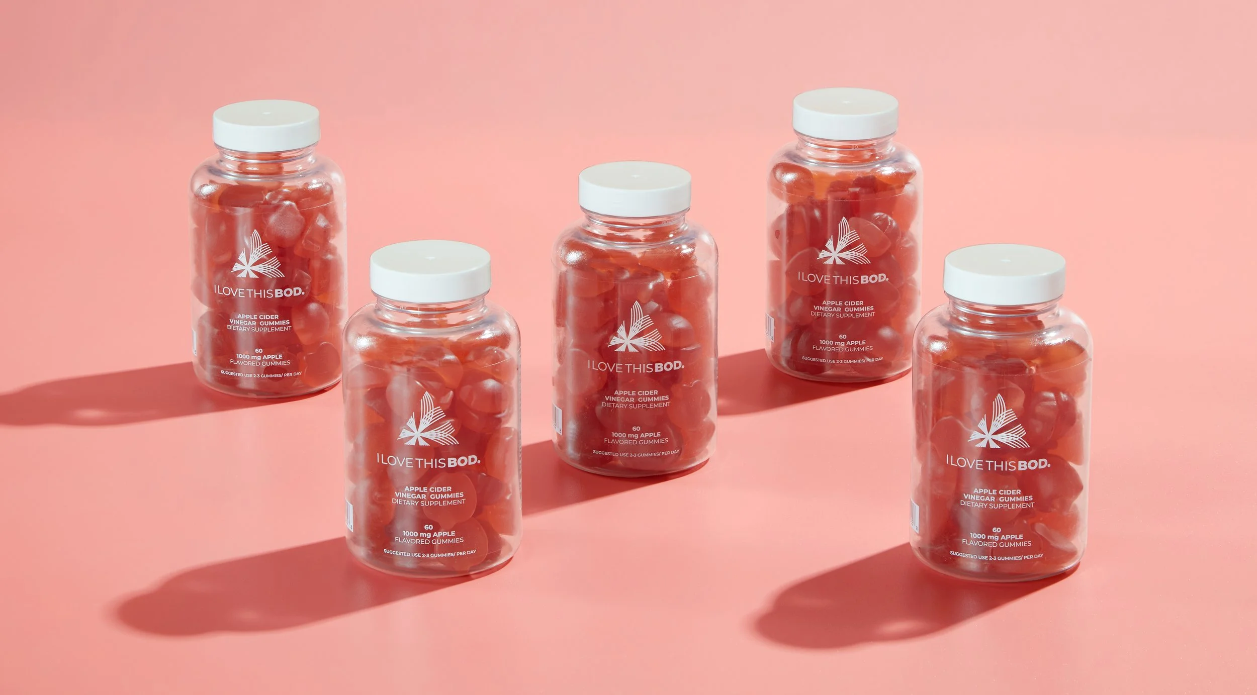 Five bottles of apple cider vinegar flavored gummies on a pink background, with each bottle containing red gummy candies.