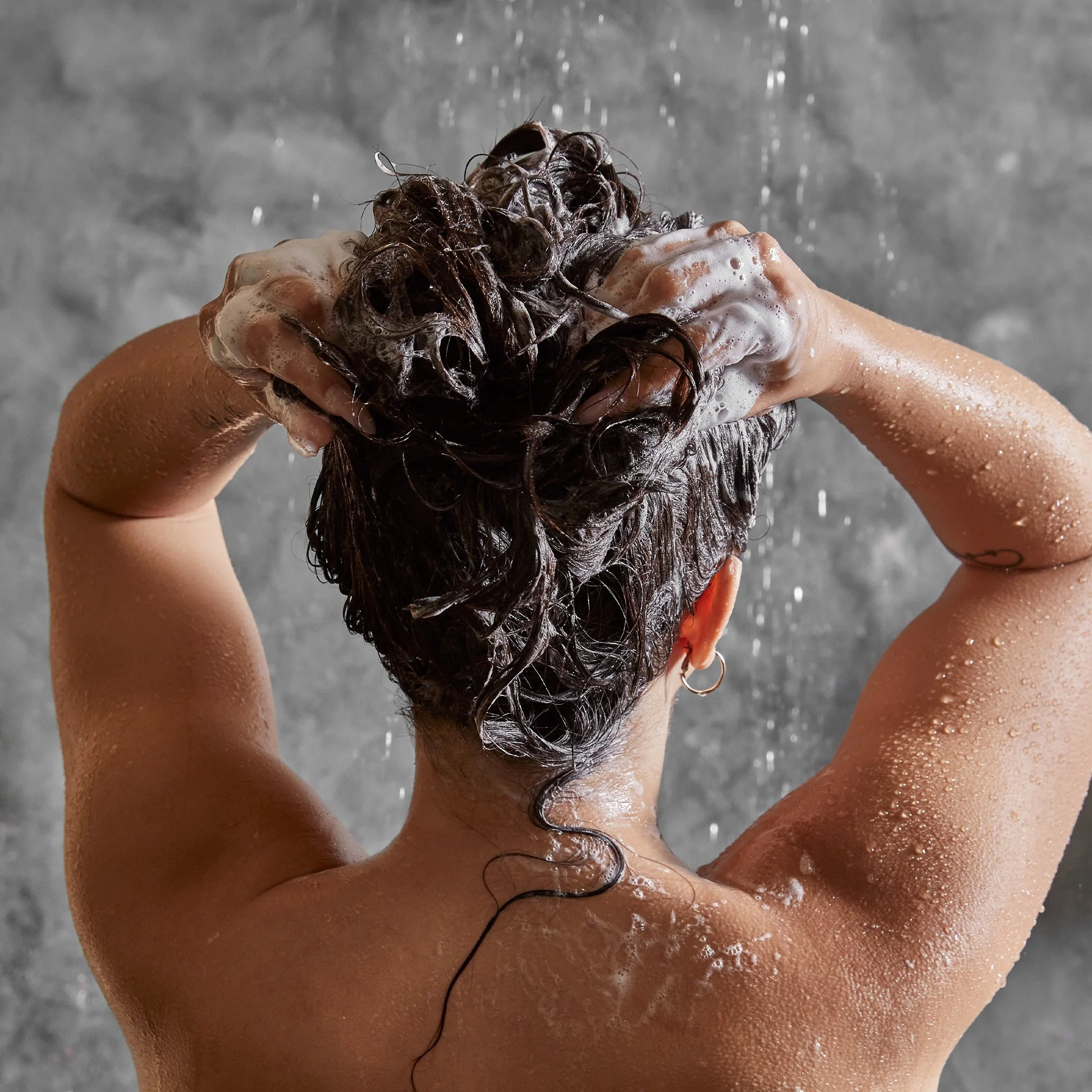 A person with short, curly dark hair, wearing a hoop earring, is washing their hair with soap and water in the shower, facing away from the camera, with water and soap suds visible on their hair and body.