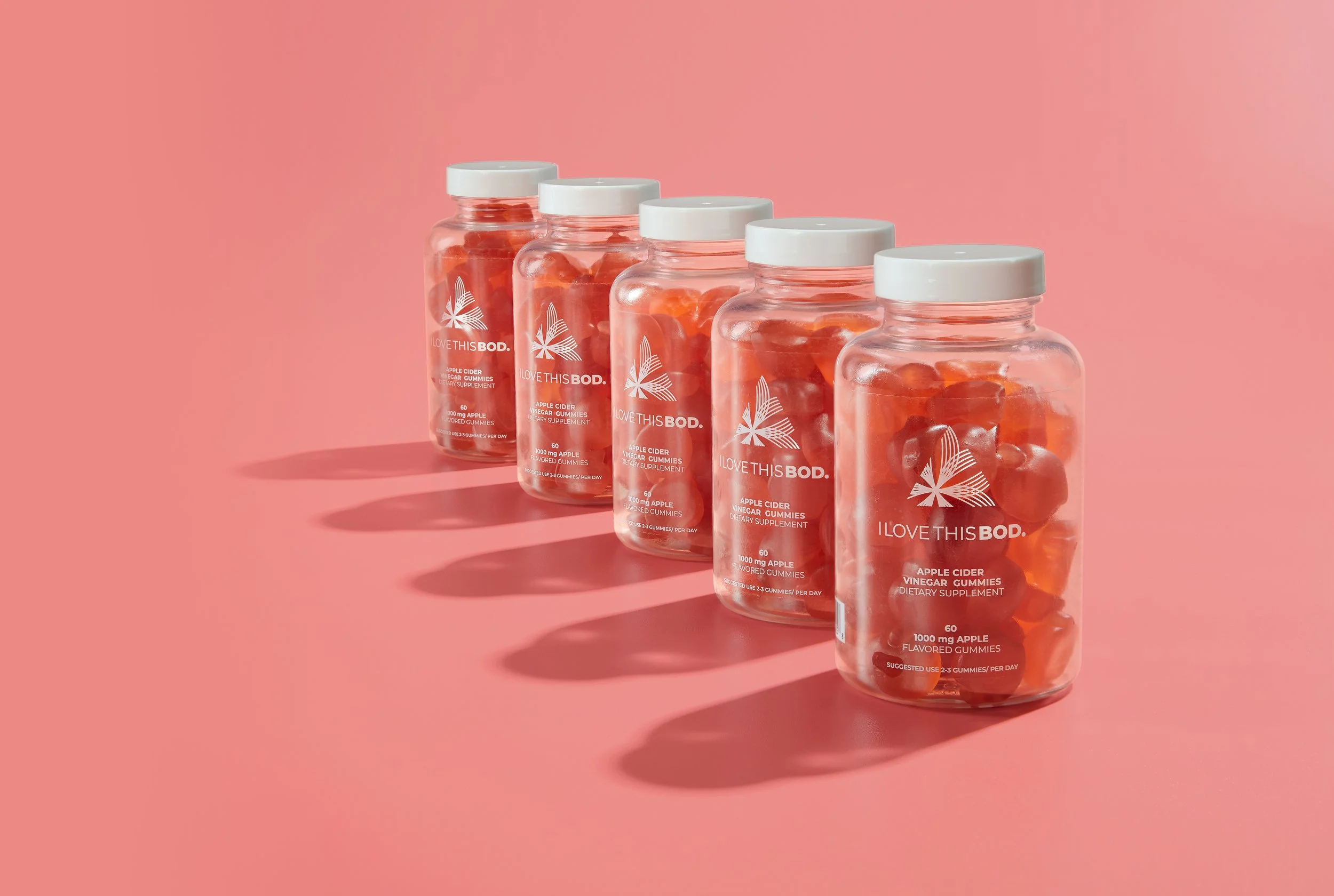 Five ILOVETHISBOD. bottles of apple cider vinegar gummies lined up on a pink surface, casting shadows.