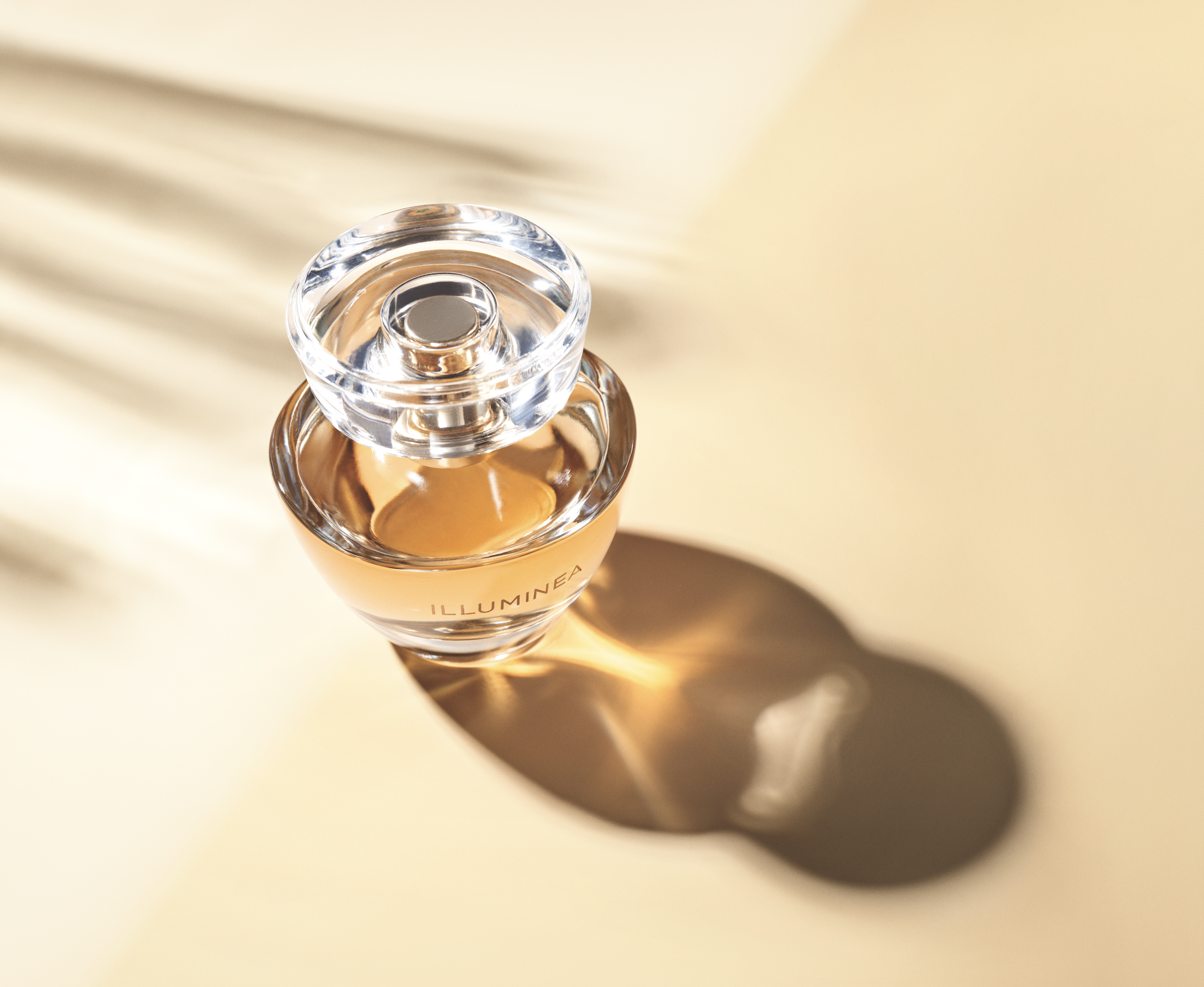 A perfume bottle labeled 'ILLUMINEA' with a clear glass cap, casting a shadow on a light-colored surface with shadows from nearby blinds.