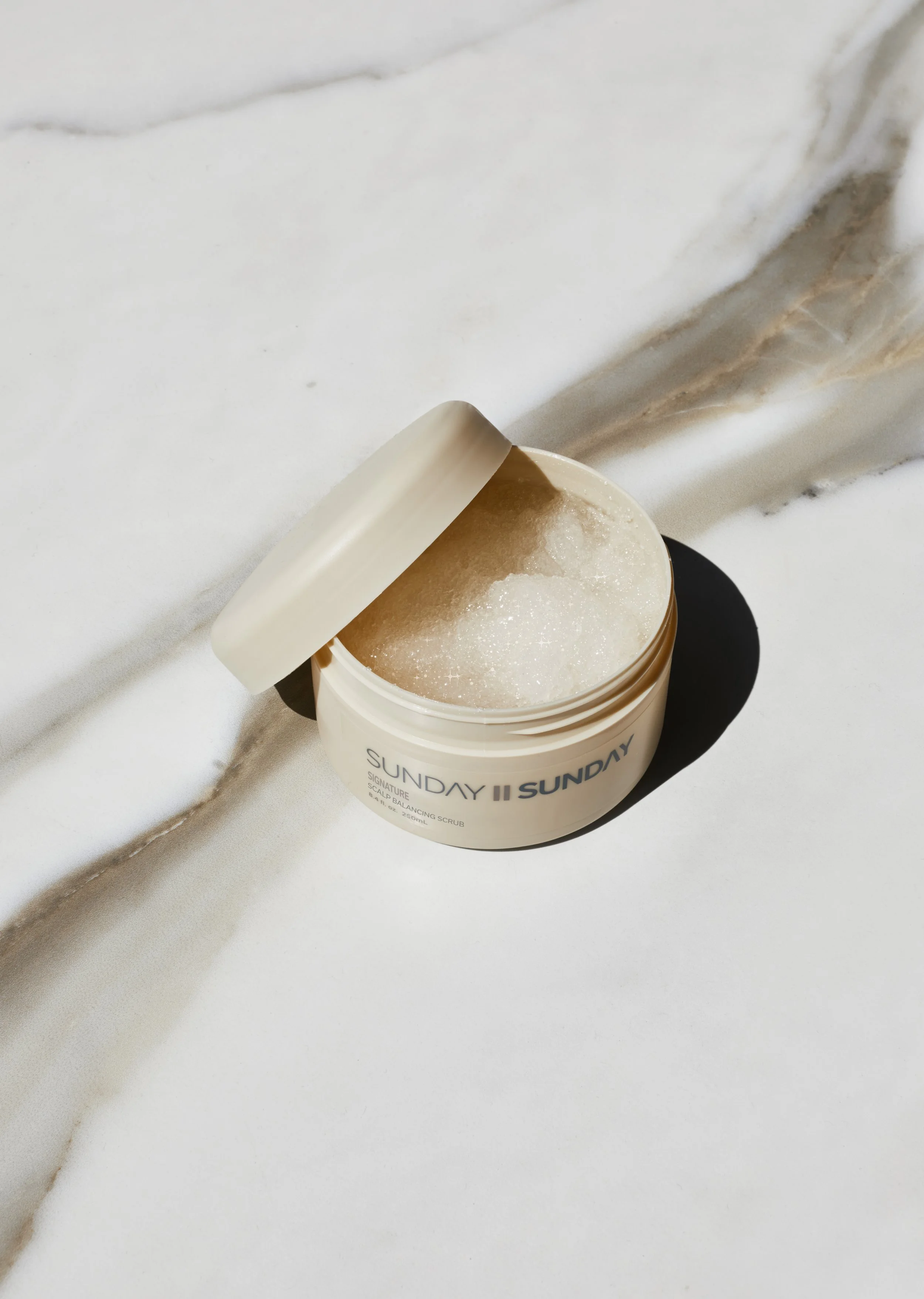 Open container of Sunday II signature salt balancing scrub on white surface with marble pattern, partially tipped open showing white exfoliating scrub inside.