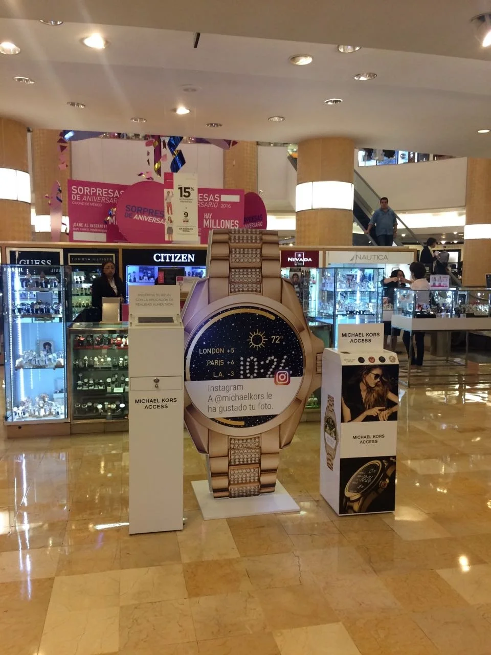 Display stand in a mall showing a large digital watch face with a gold band, surrounded by jewelry stores and other retail displays.