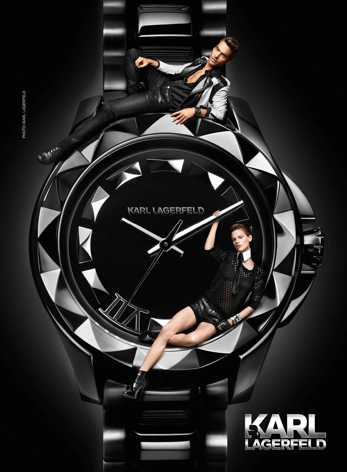 Two models are positioned on and around a large, black and silver Karl Lagerfeld watch with a black background. One man is lounging on top of the watch, leaning back with one arm and legs crossed. The woman is sitting inside the watch face, with one 