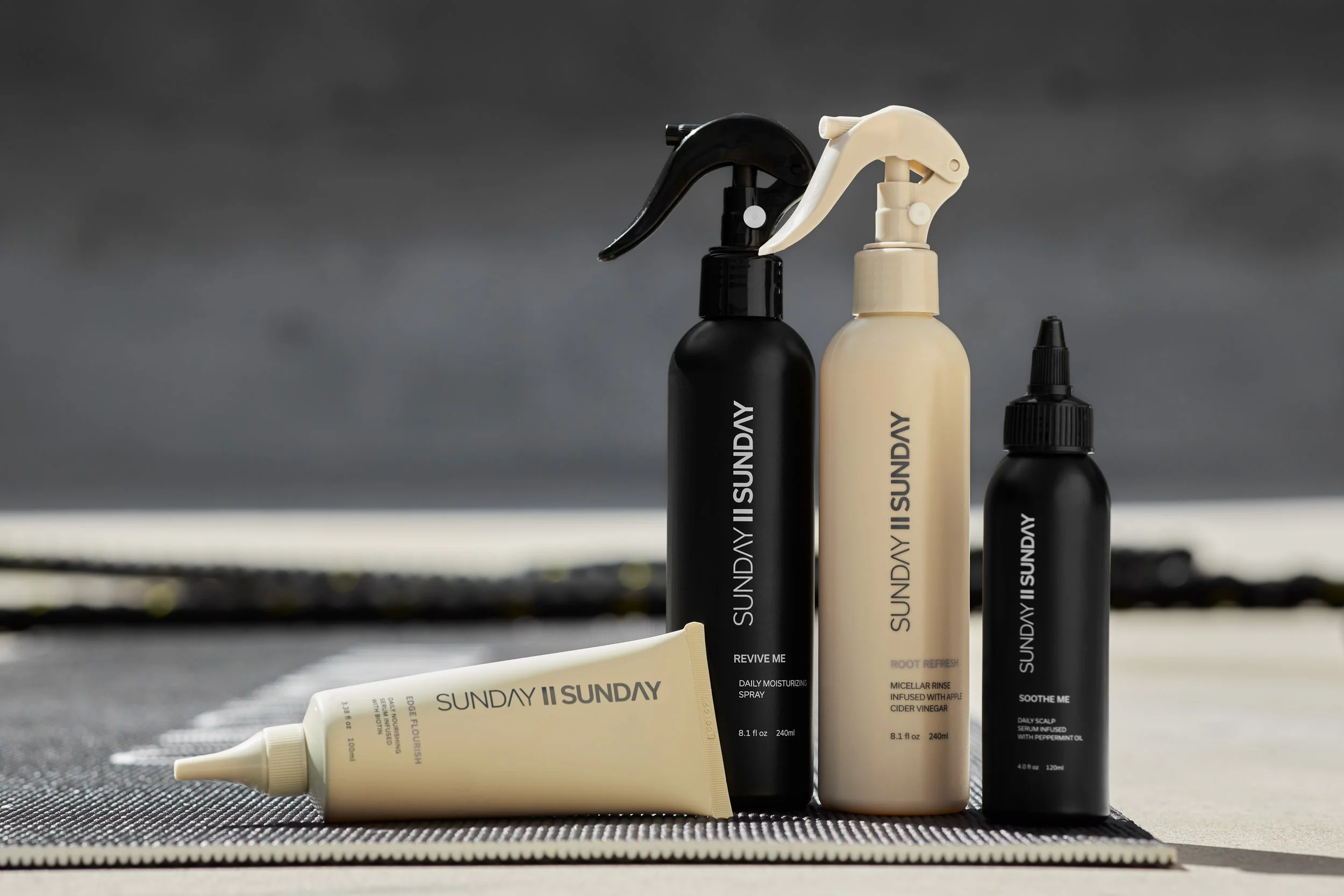 Collection of Sunday II Sunday haircare products with black and beige packaging on a textured mat against a cloudy sky background.