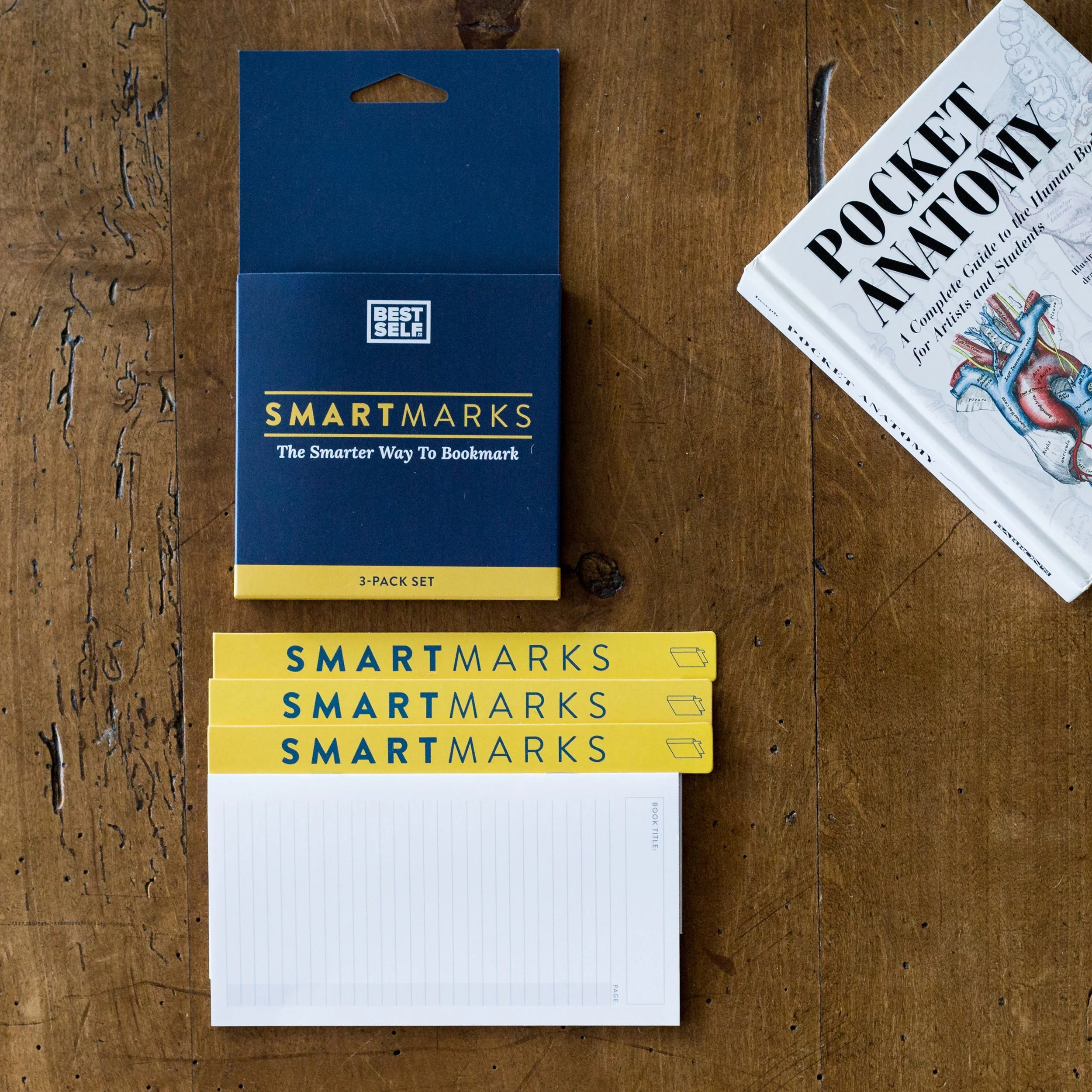 A set of three yellow and blue SmartMarks bookmarks, a matching blue box, a white notepad, and a book titled 'Pocket Anatomy' on a wooden surface.