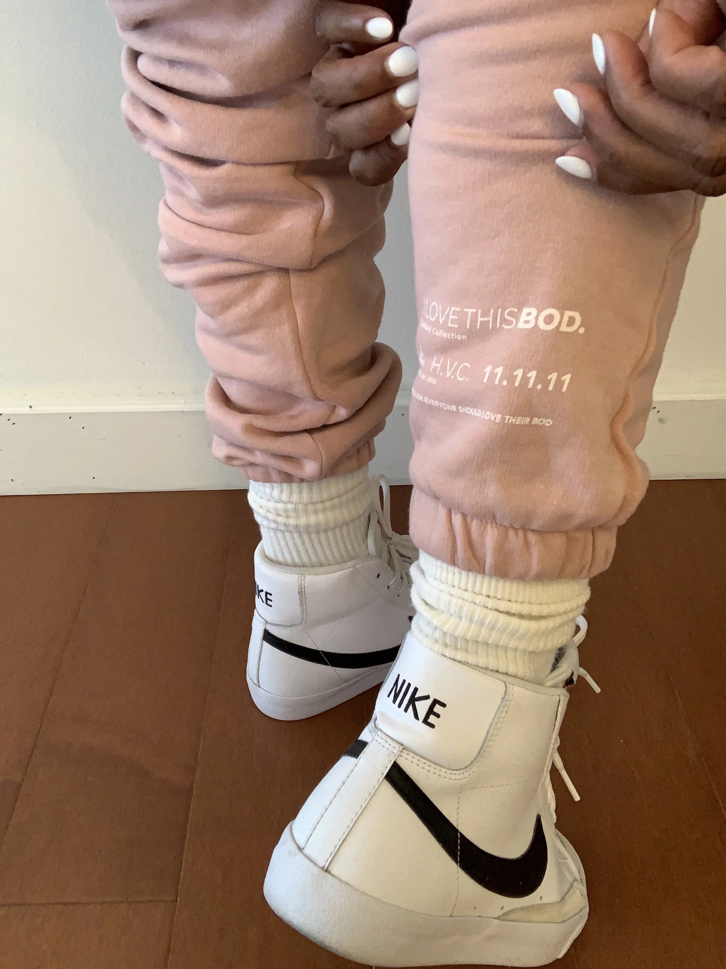 Person wearing pink sweatpants with text, white Nike sneakers, and cream-colored socks, standing on a wooden floor.