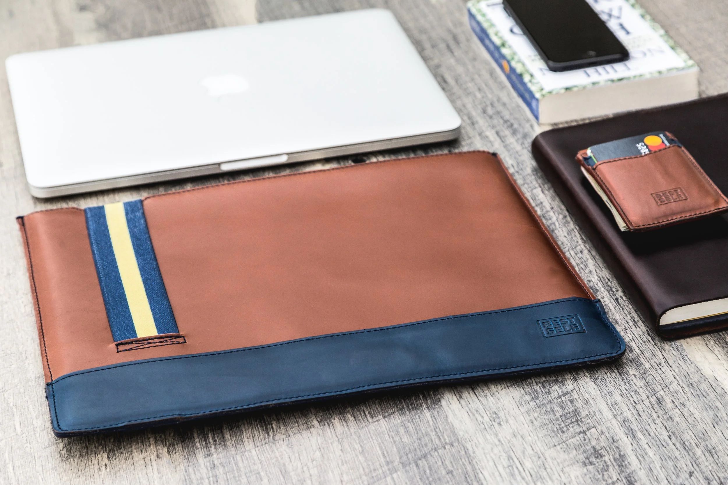 Desk with a closed laptop, a stack of books with a phone on top, a brown wallet with cards, a black notebook, a brown leather file organizer, and a colorful band on the organizer.