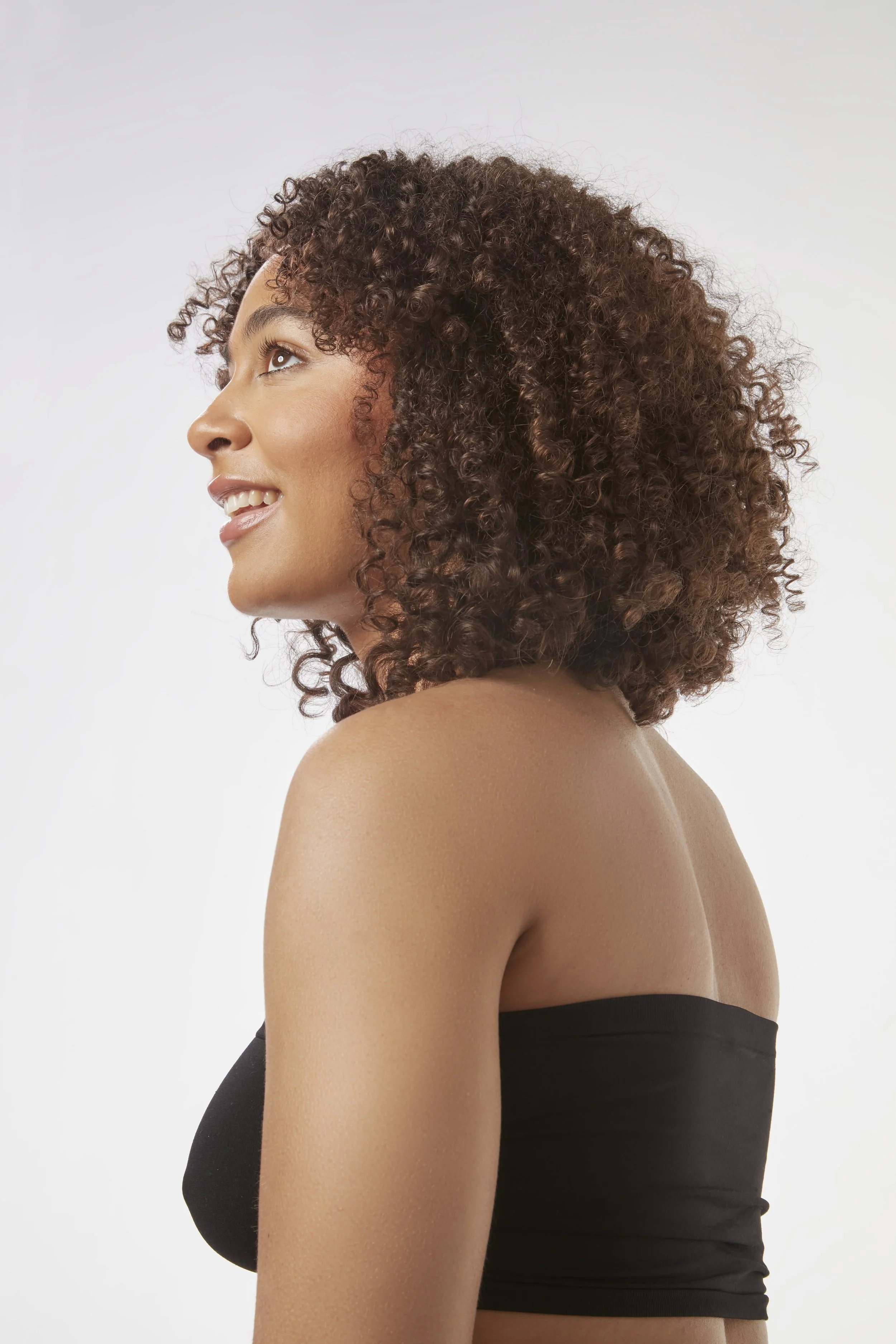 Side profile of a woman with curly brown hair smiling, wearing a black tube top, against a plain white background.