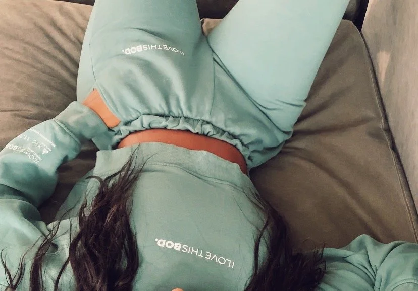 A woman is lying on a gray couch, wearing a matching mint green sweatshirt and shorts with the text 'LOVETHISBOT' on both, and has long dark hair.