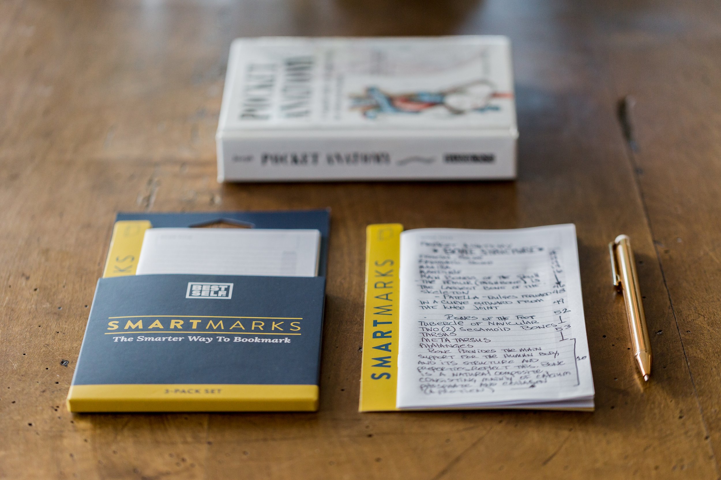 A wooden table with a box of SmartMarks bookmarks by Best Self, a notepad with handwritten notes, and a gold pen placed beside the notepad.