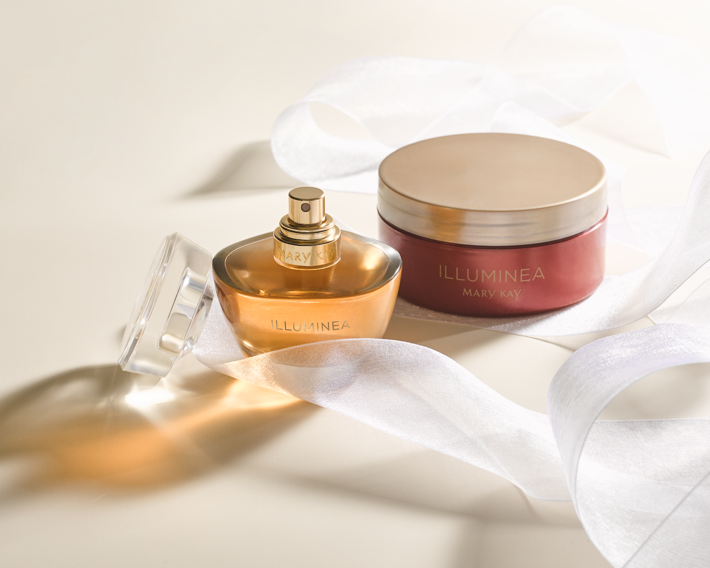 A Mary Kay Illuminea perfume bottle and a matching pink and gold container with white ribbon on a white surface.