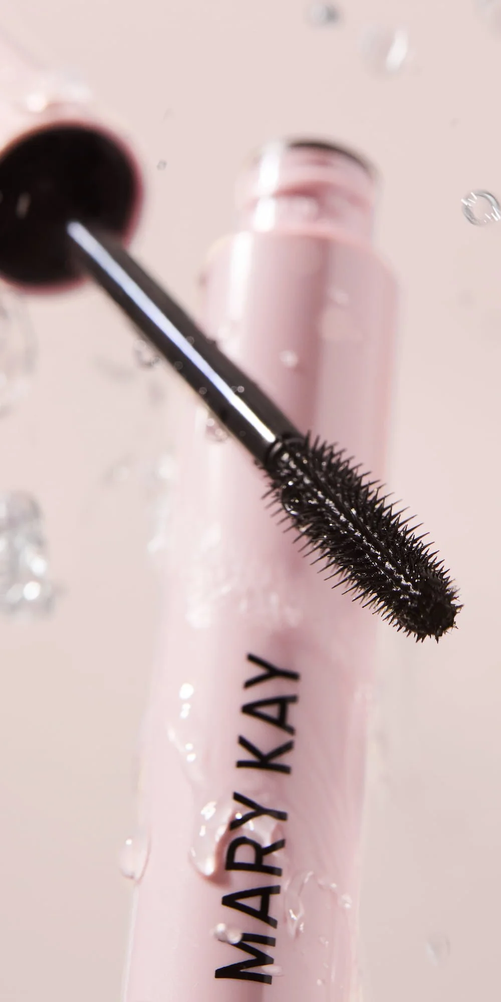 Close-up of a black mascara wand resting on a pink lip gloss container with water droplets on its surface, labeled "MIARY KAY".
