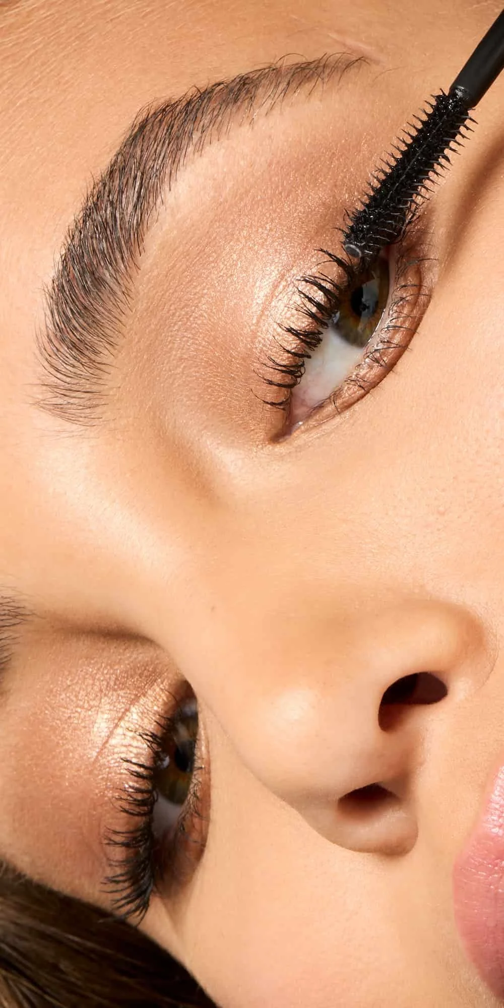 Close-up of a woman's eye with mascara wand applying makeup, showcasing well-groomed eyebrows and shimmering eyeshadow.